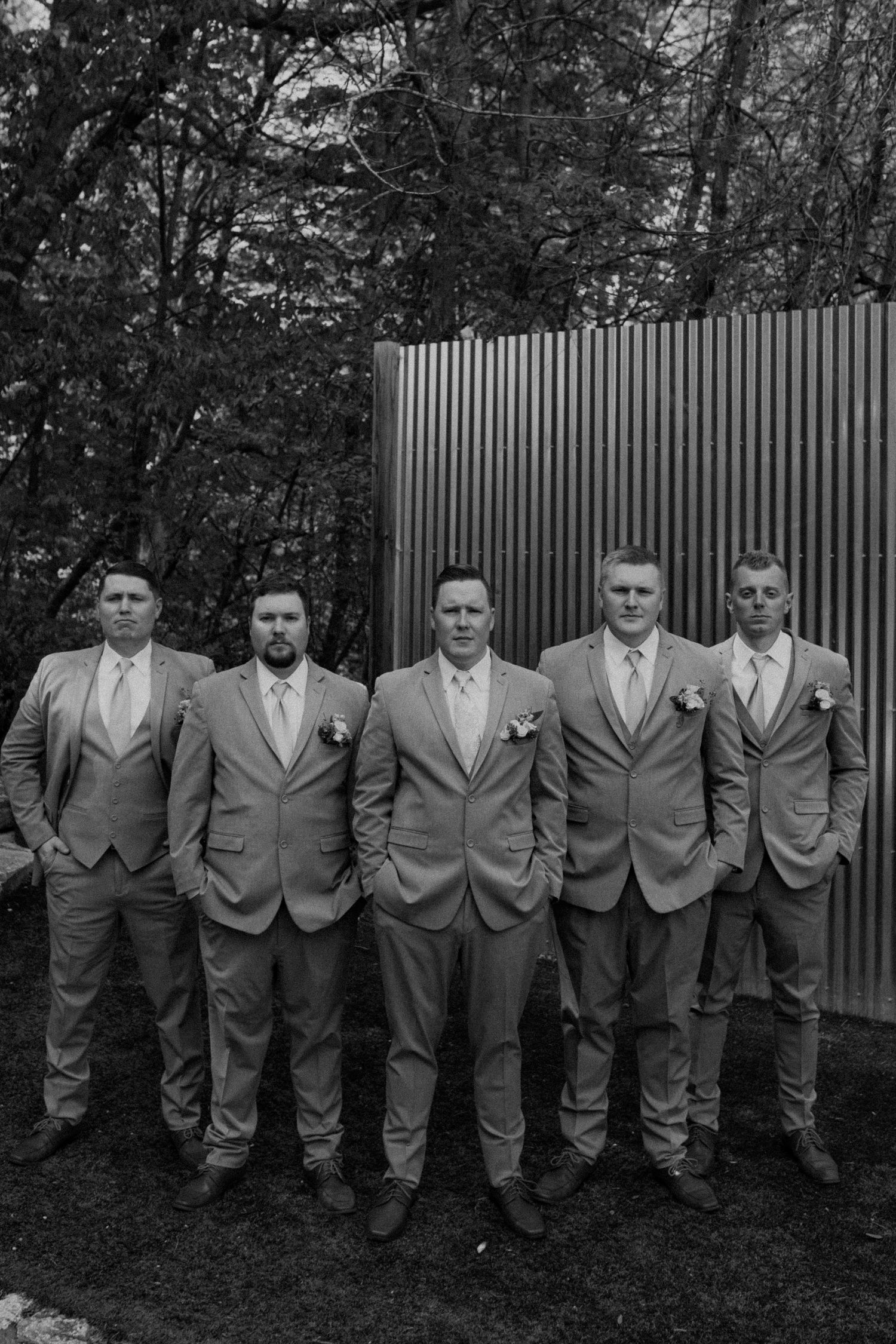 A group of groomsmen in matching gray suits and vests pose together against a wooden fence backdrop.