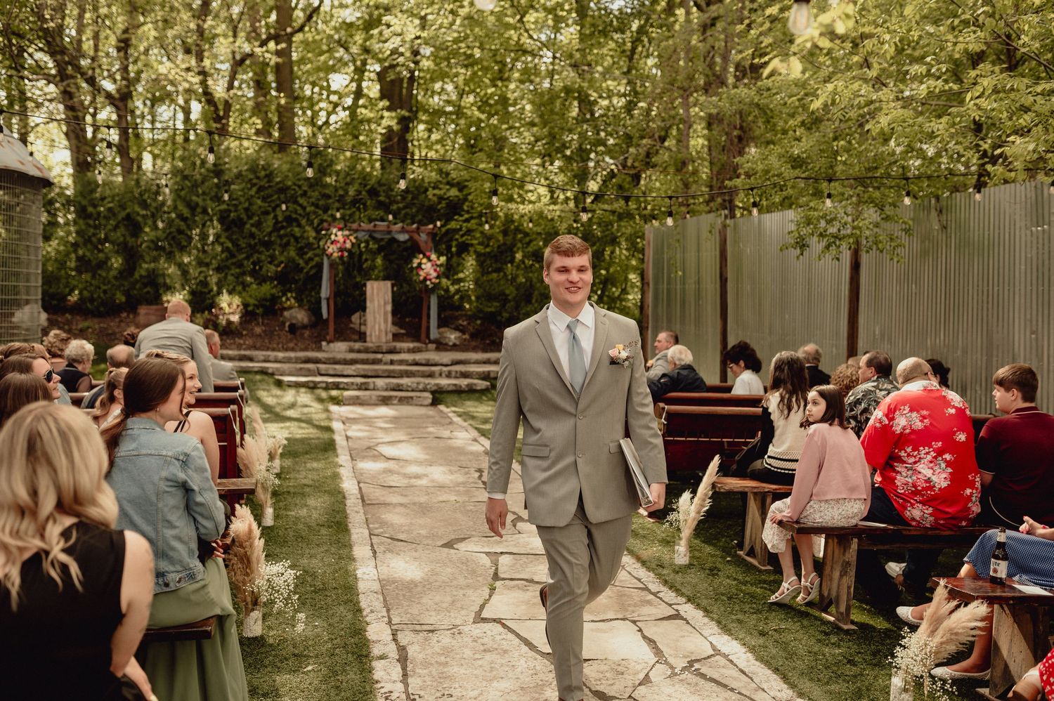 A groomsman walks down an outdoor garden path between seated wedding guests during a summer ceremony.