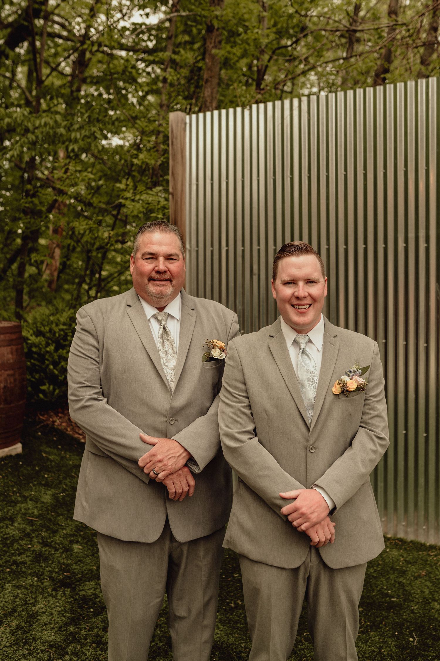 Two groomsmen in matching tan suits and boutonnières pose together in front of a garden fence.