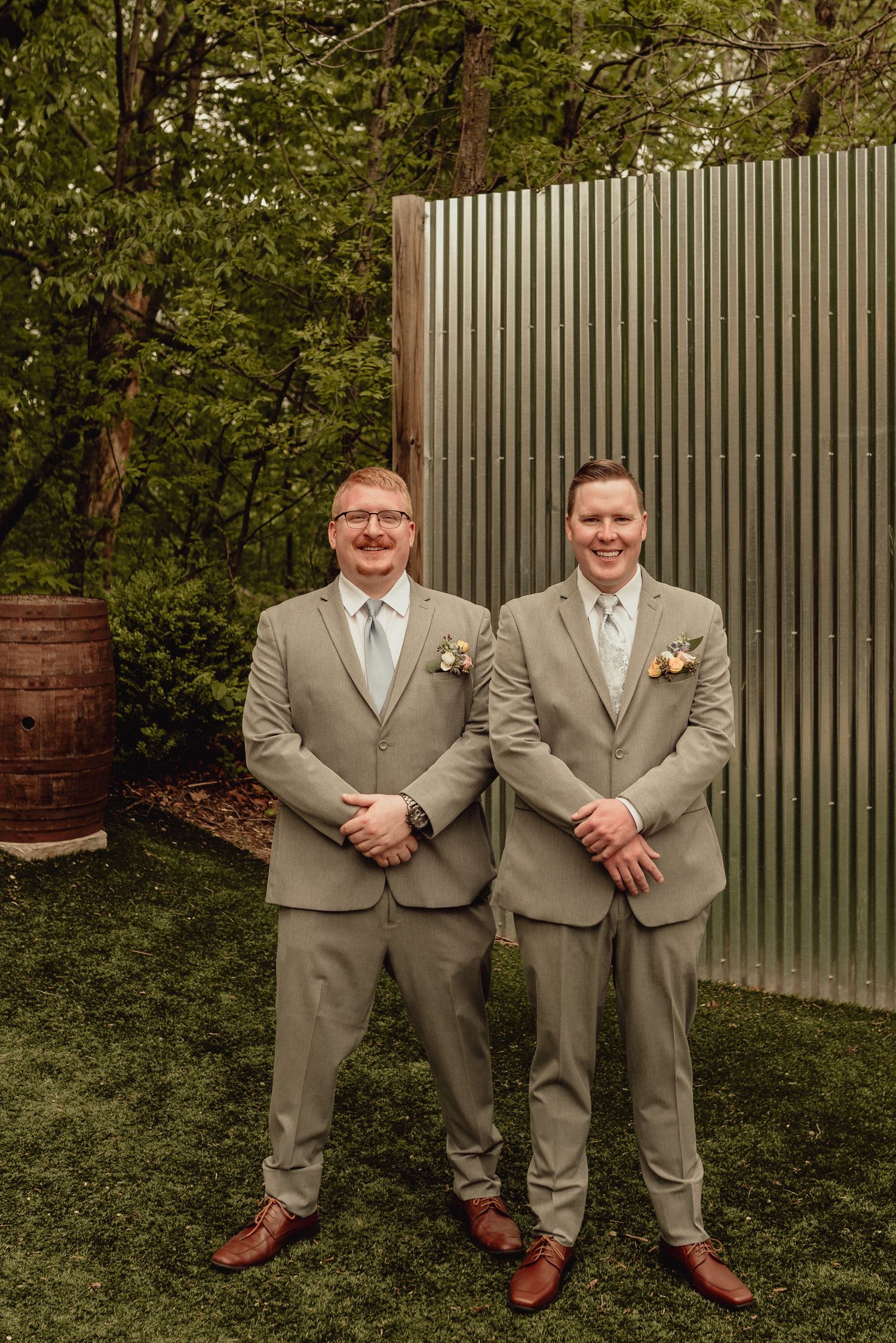 Two people in tan suits and brown shoes stand together in a garden setting with a fence backdrop.