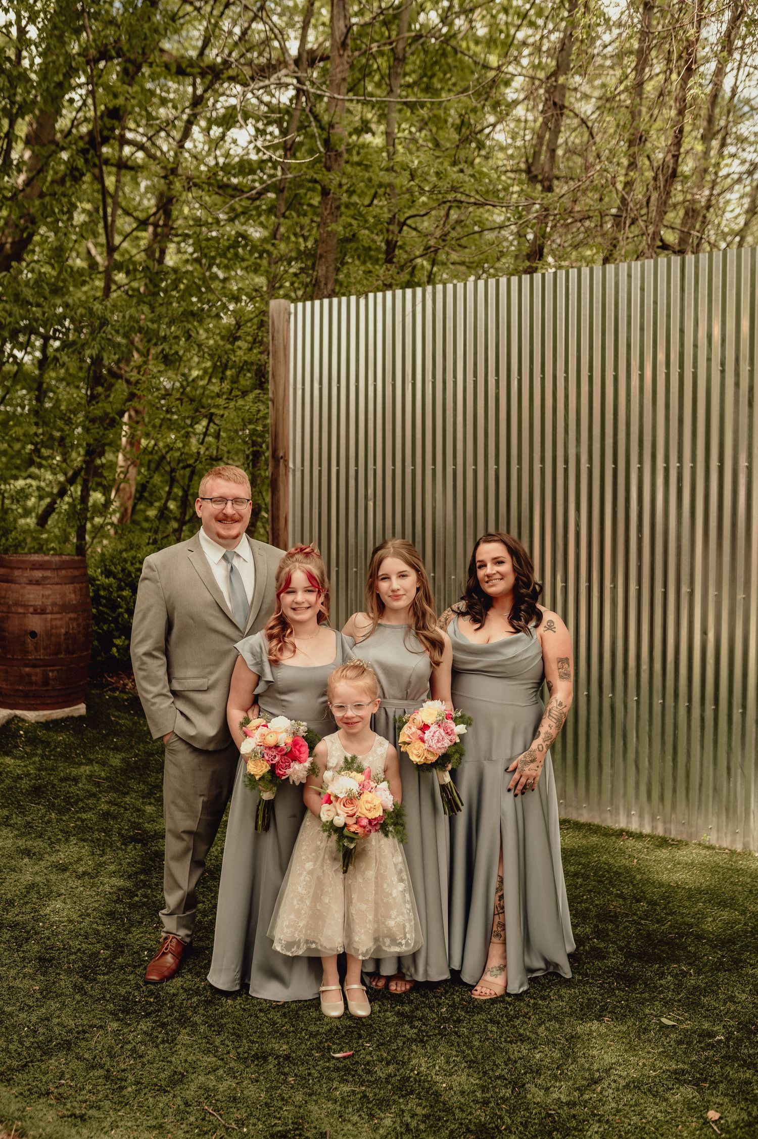Wedding party in gray attire poses against a corrugated metal wall and greenery backdrop at outdoor venue.