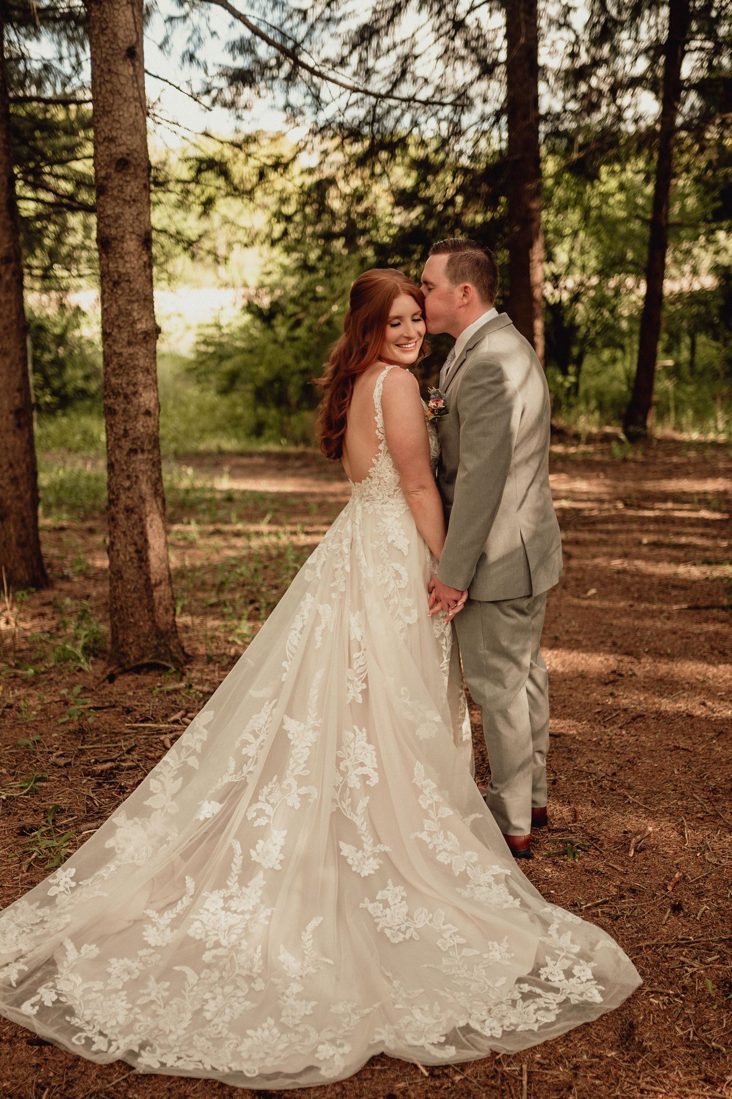 A romantic wedding couple embraces in a serene pine forest setting as sunlight filters through the trees.