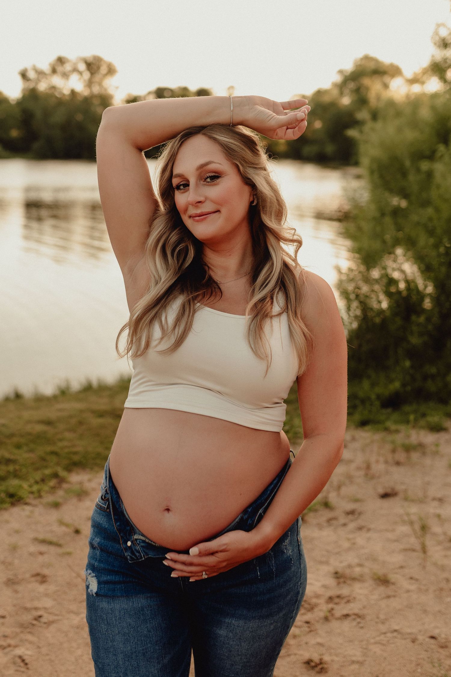 Maternity photoshoot at sunset by a lake with a white crop top and distressed jeans.