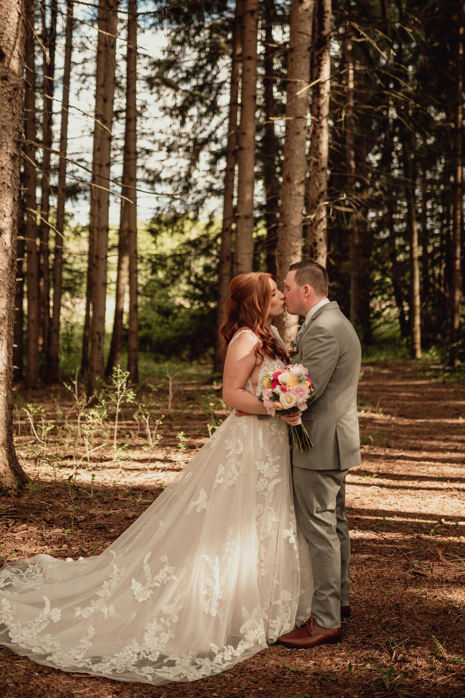 Romantic wedding couple shares intimate moments in a sunlit forest setting with a flowing white dress and gray suit.