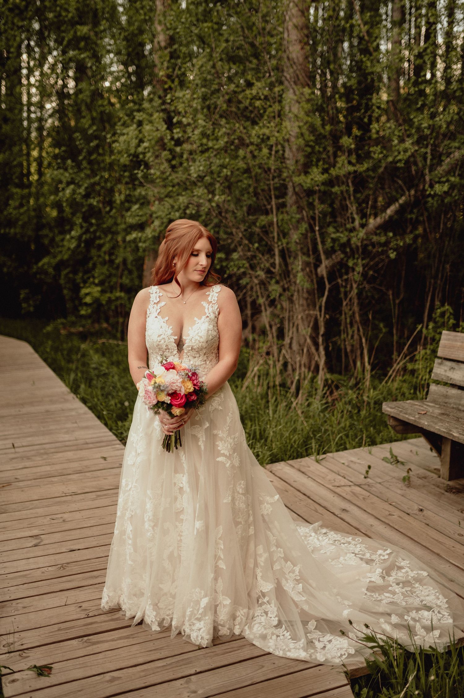 A bride in a lace wedding dress sits gracefully on a wooden boardwalk surrounded by lush forest scenery.
