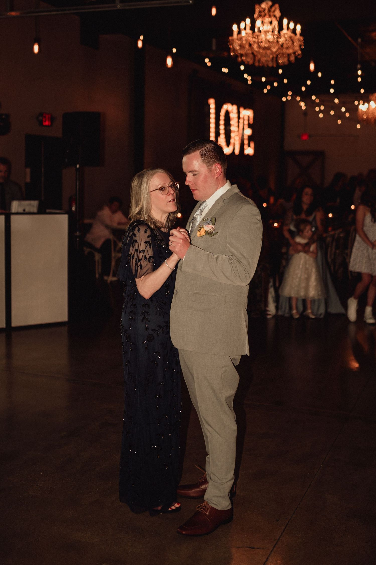 A couple shares an intimate dance moment under a chandelier and string lights at an indoor wedding reception.