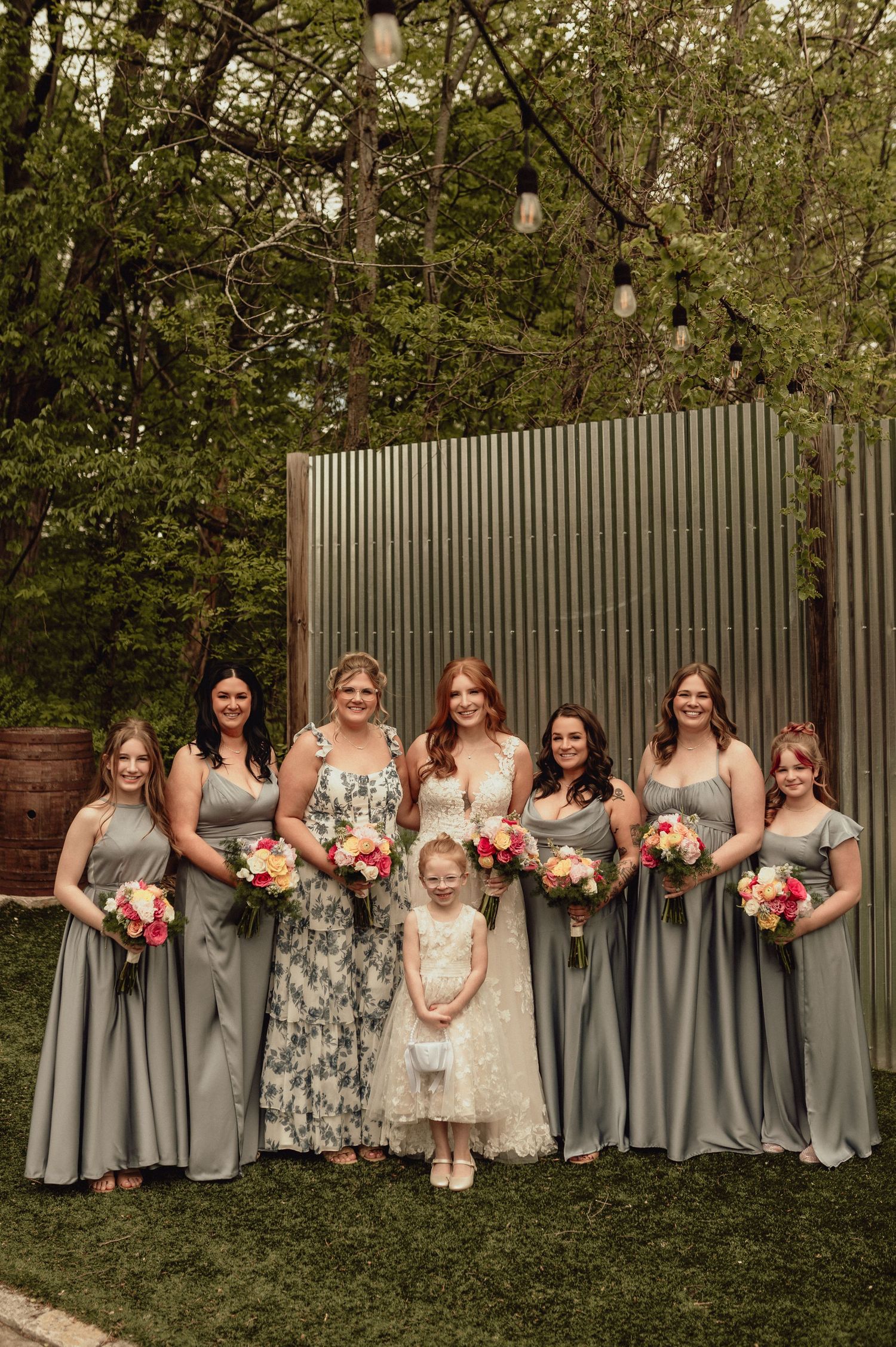 Wedding party wearing gray bridesmaid dresses and floral gowns pose together on grass with a gray fence backdrop.