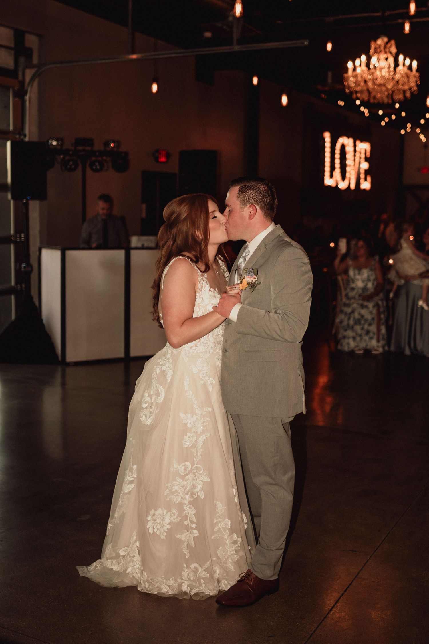 Newlyweds share a romantic dance together in a dimly lit reception venue with chandeliers overhead.