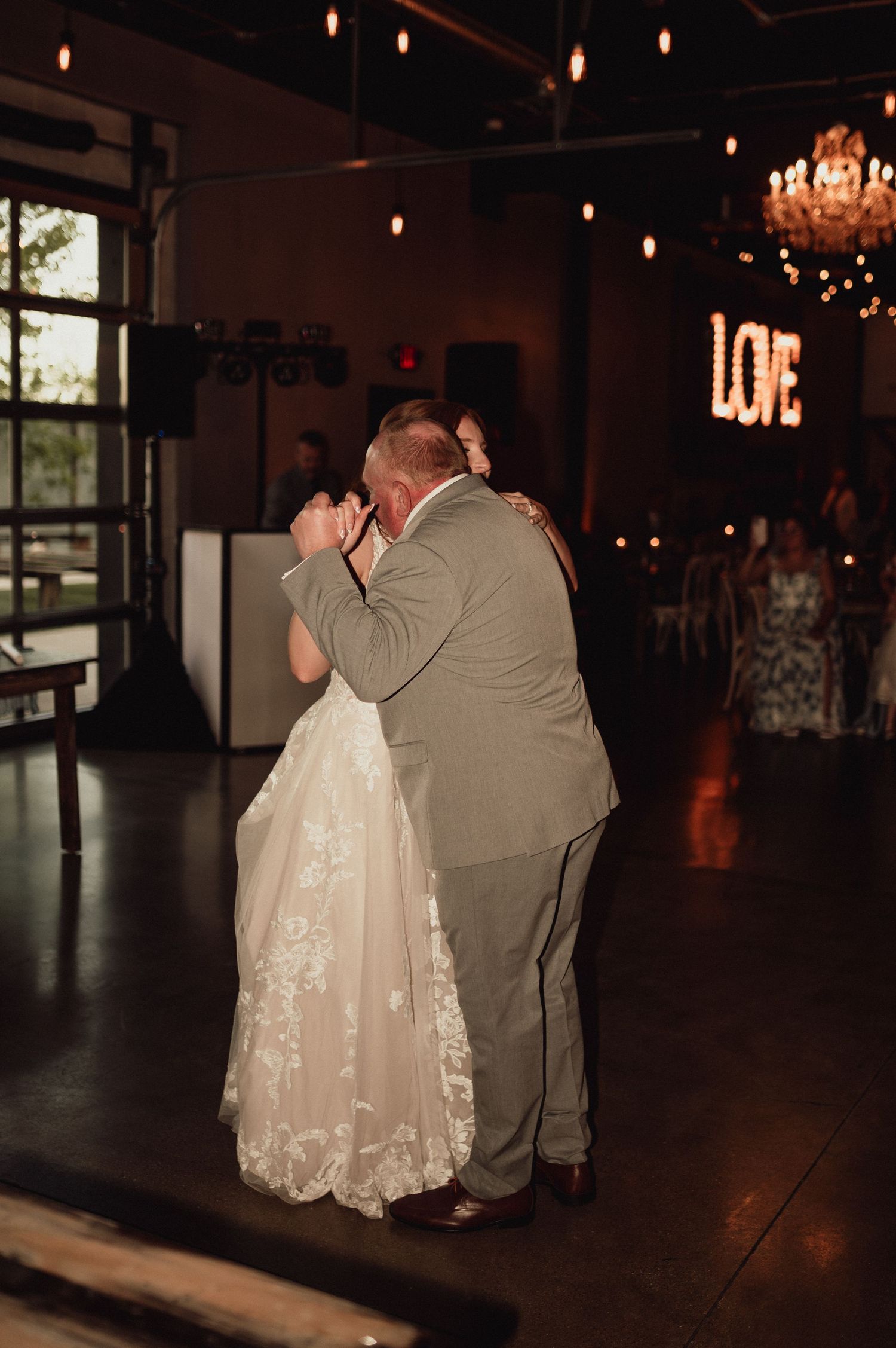 A couple shares a tender dance moment in a dimly lit reception hall with chandeliers and string lights overhead.