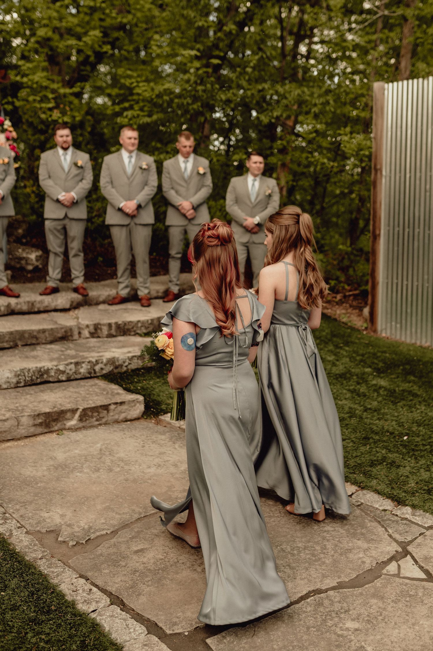 Wedding party members in gray suits and dresses walk along a stone path surrounded by lush greenery.
