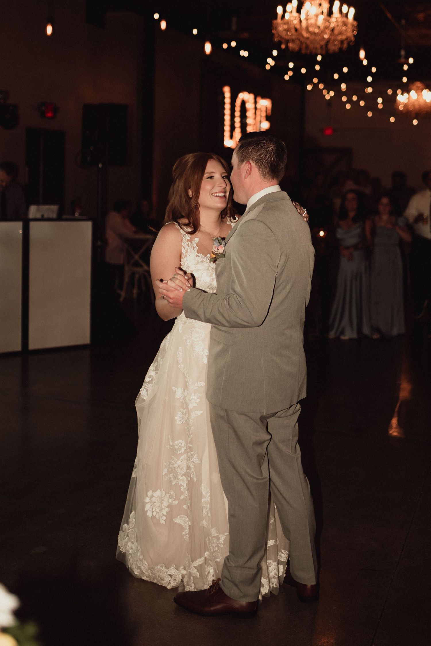 A couple shares a romantic dance together at their wedding reception under glowing chandeliers.