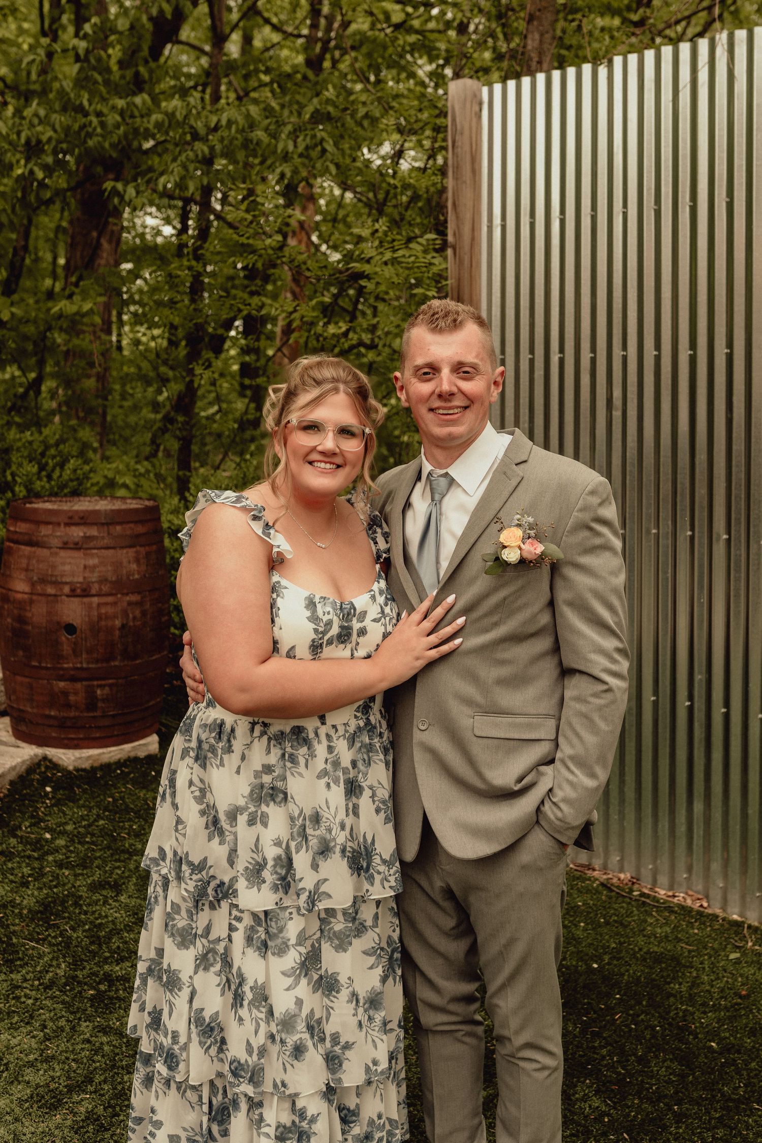 A couple poses outdoors at a rustic wedding venue with a wooden barrel and metal wall backdrop in natural lighting.