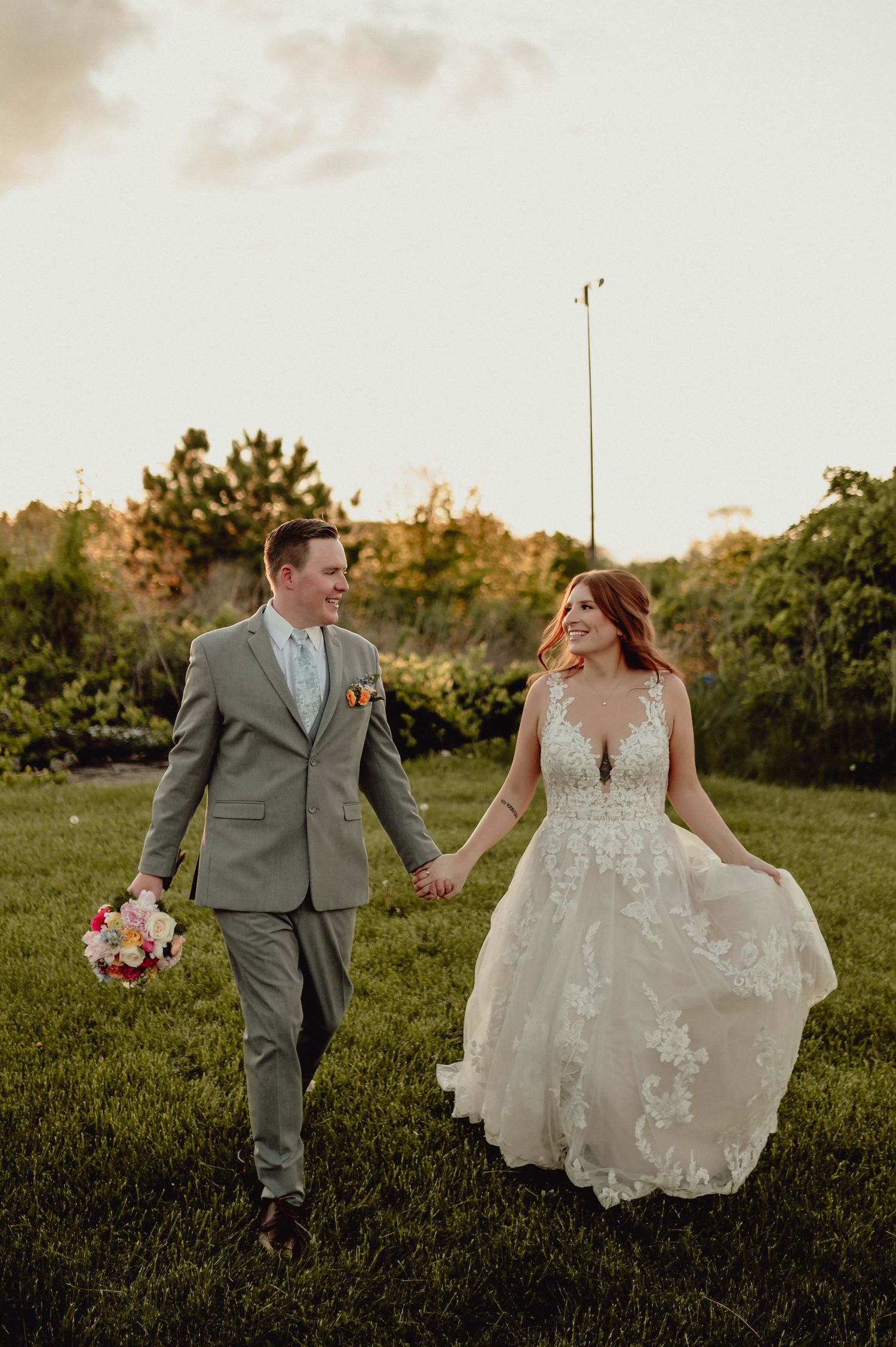 Newlywed couple walks hand in hand through a sunlit meadow at sunset wearing sage green suit and lace wedding gown.