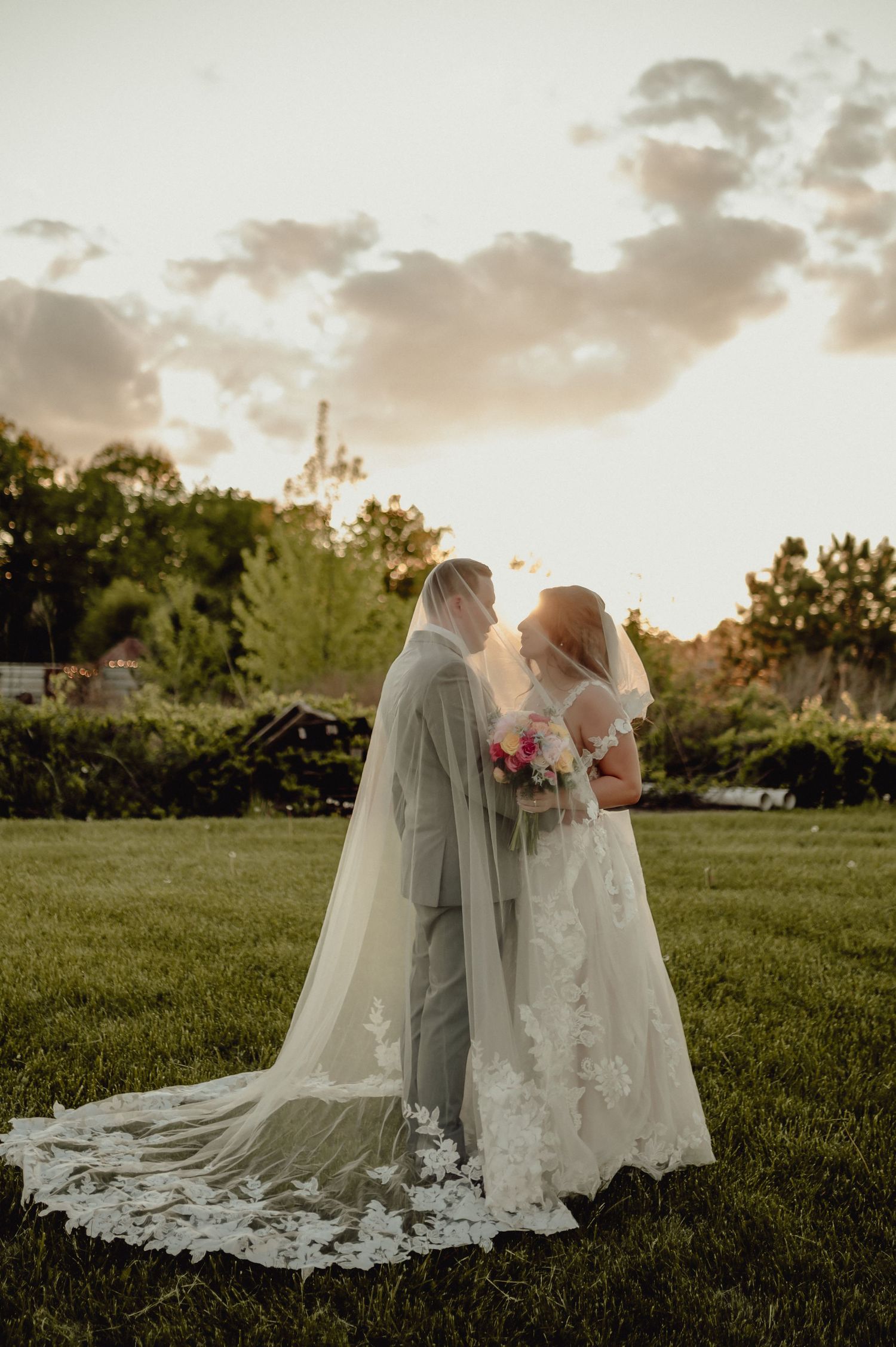 A couple in wedding attire embraces on a grassy field during golden hour sunset.