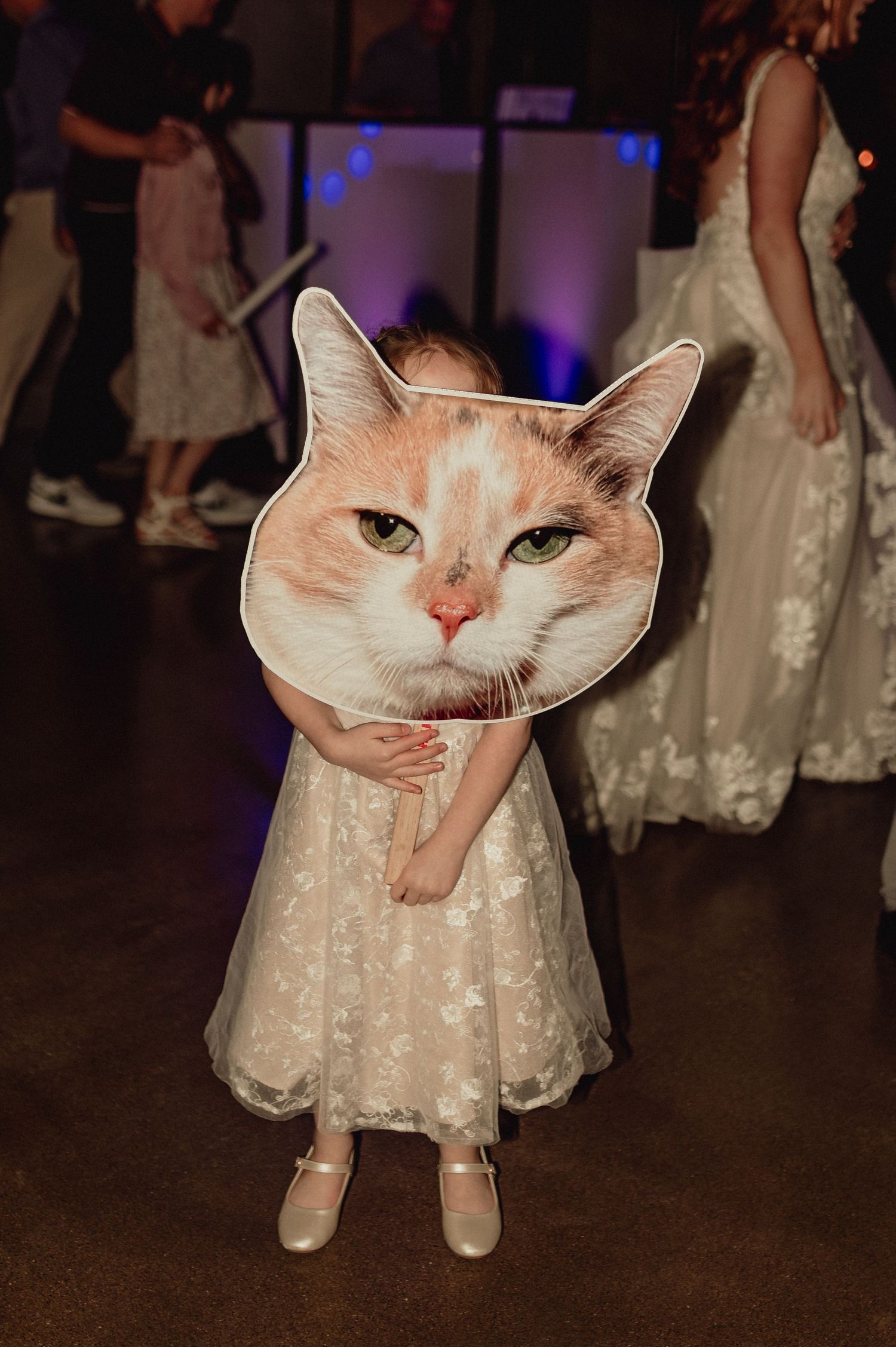 Child in white lace dress holds large cat face cutout at wedding reception with purple lighting in background.