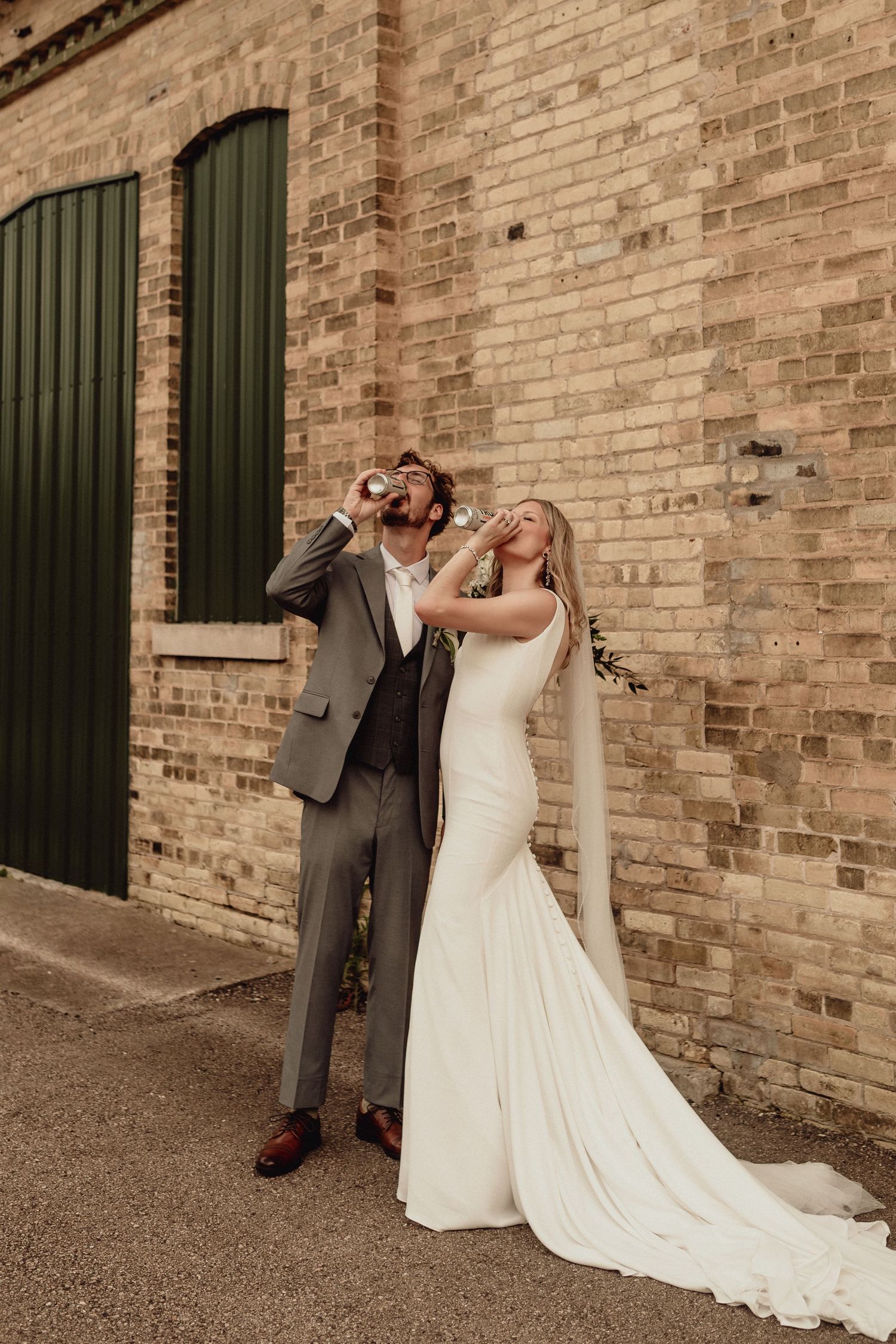 Playful wedding couple taking shots together against a brick wall.