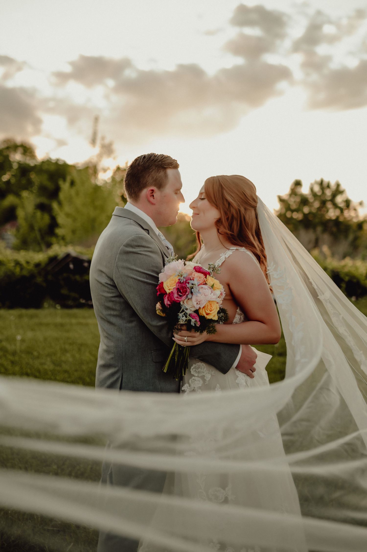Romantic wedding couple shares a tender kiss at sunset with bride's flowing veil creating an ethereal atmosphere.