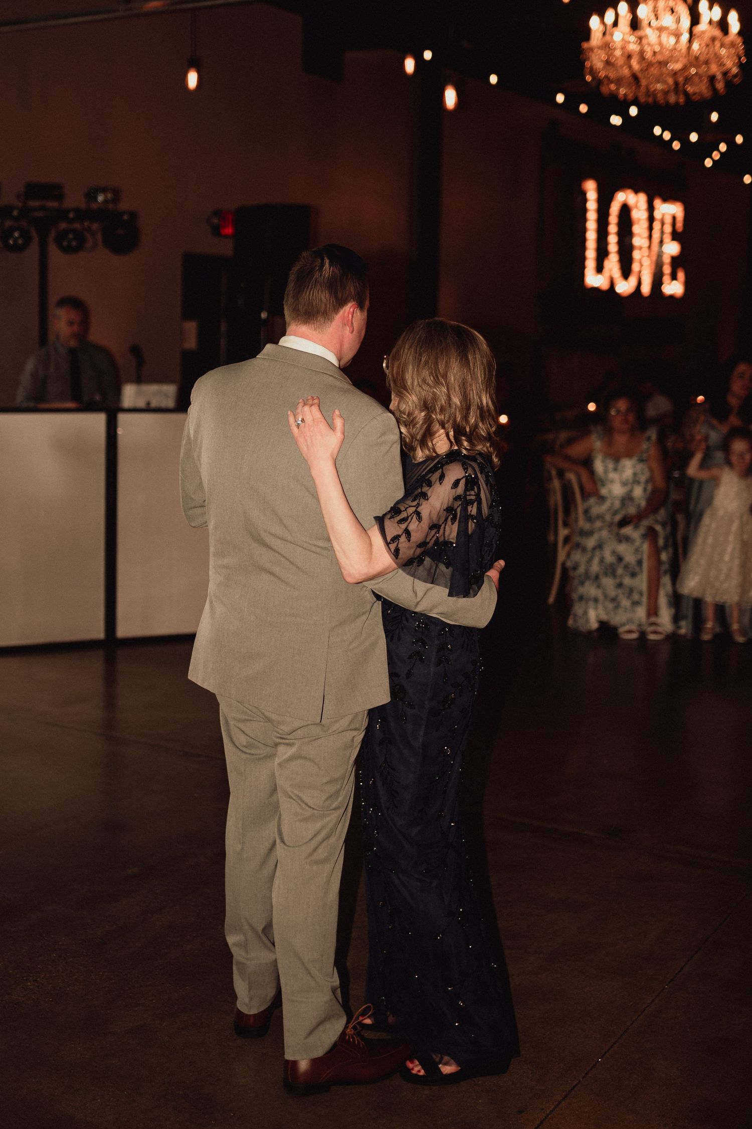 A couple dances together at a wedding reception under a glowing chandelier and LOVE sign.