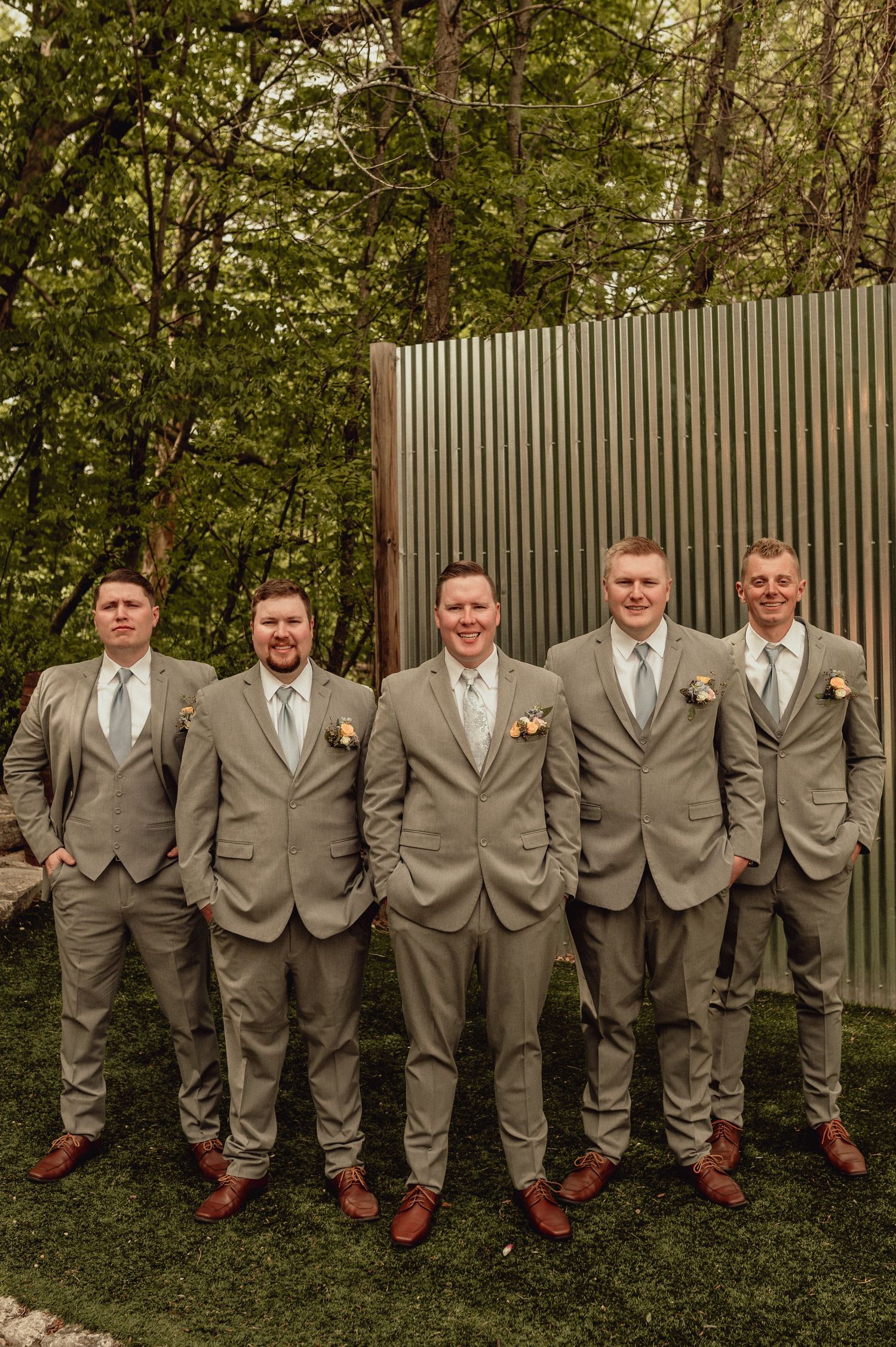 Groomsmen in matching tan suits and brown shoes pose together on grass against a corrugated wall and trees.