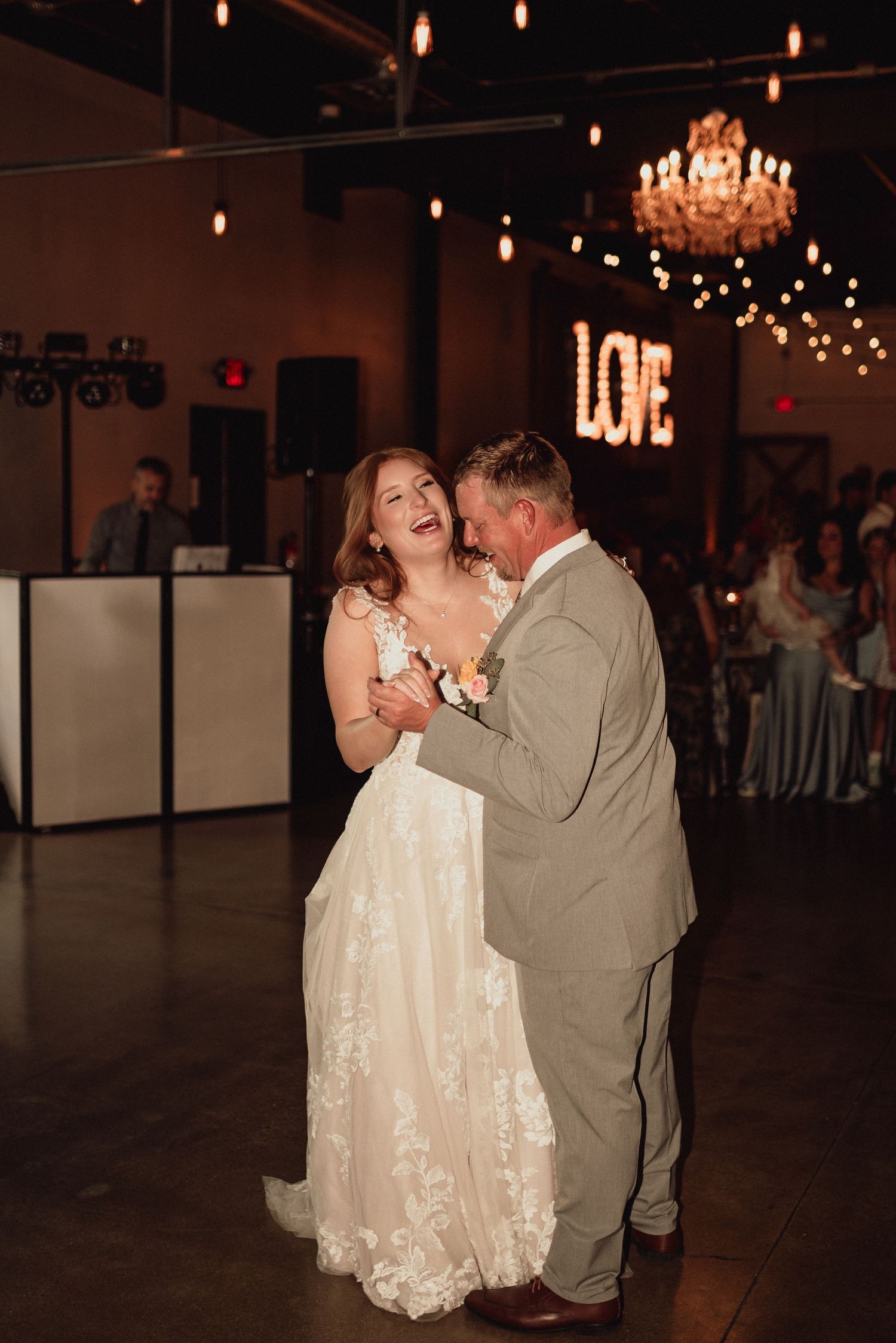 A couple shares a romantic dance moment under dimly lit chandeliers and string lights at an elegant wedding reception.