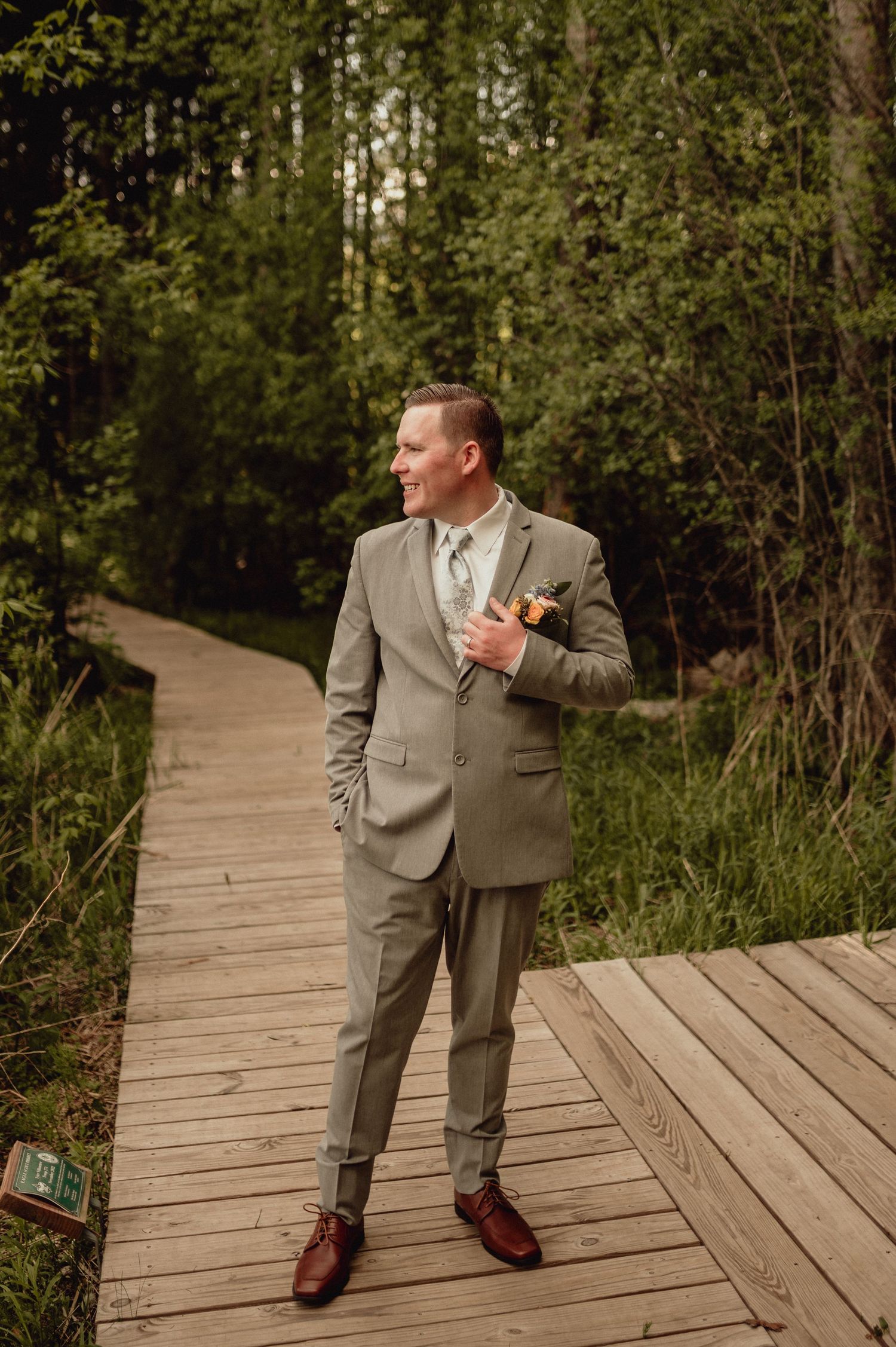 A person in a tan suit strikes playful poses on a wooden boardwalk surrounded by lush green foliage.