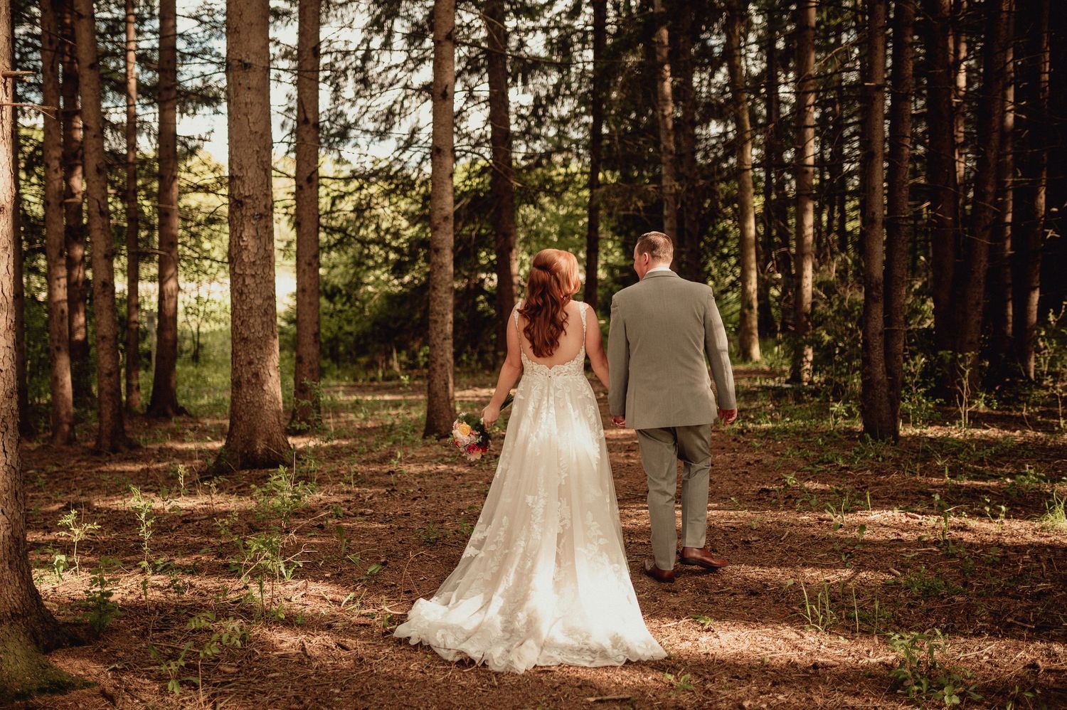 A couple in wedding attire walks hand in hand through a sunlit forest path during a romantic outdoor photo session.