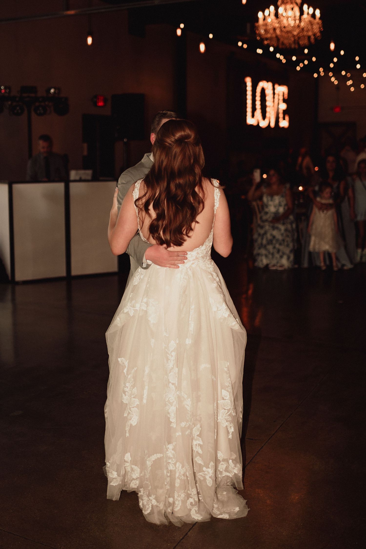 Two brides in white lace dresses dance together at their wedding reception under warm chandelier lighting.
