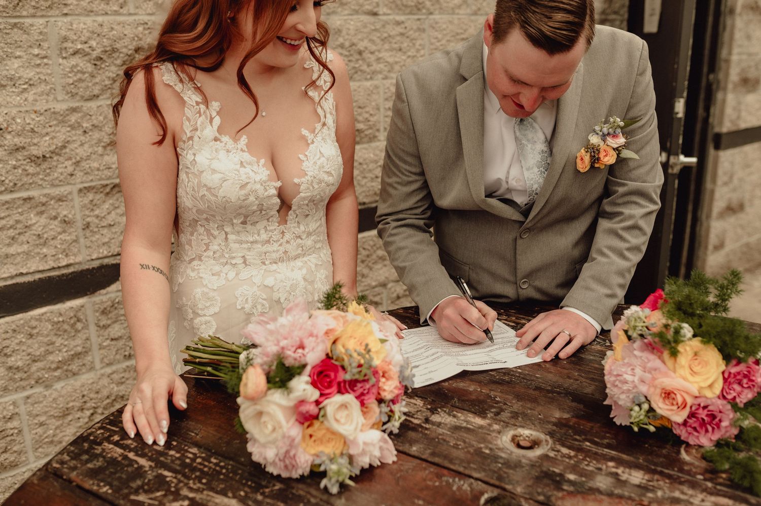 A couple signs documents at a rustic stone wall while holding a bouquet of pink and white flowers.