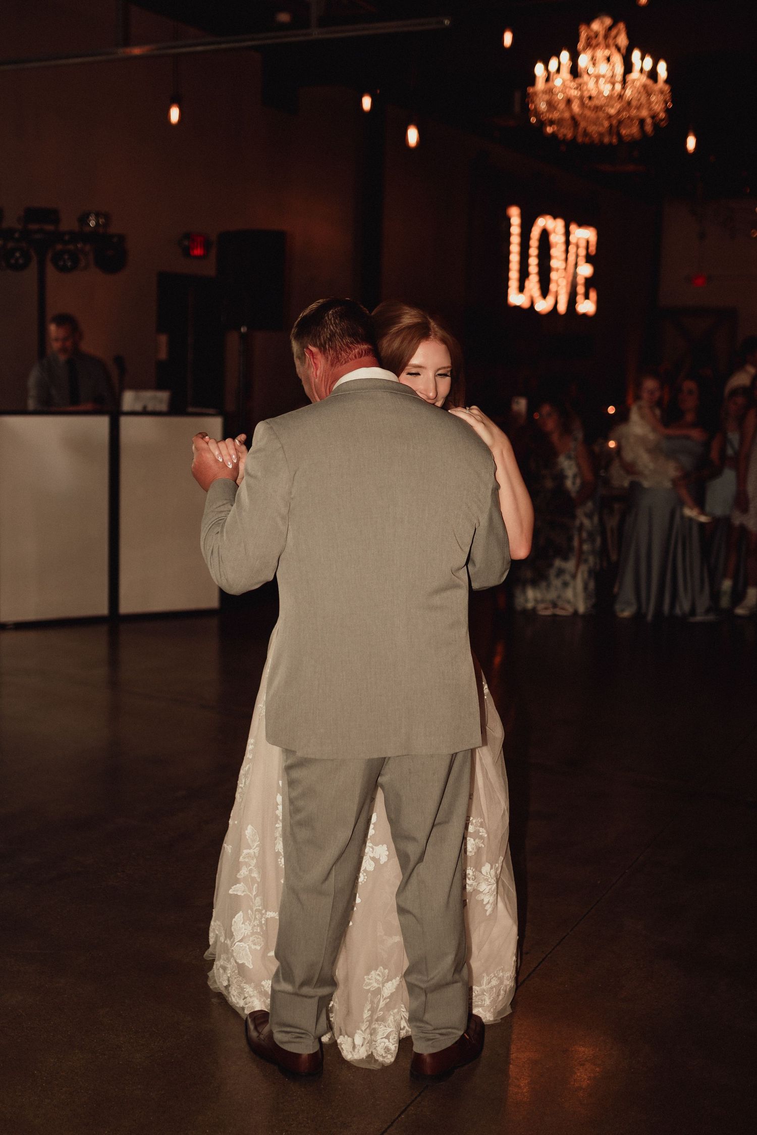 A couple dances together at a wedding reception with a chandelier visible in the dark background.