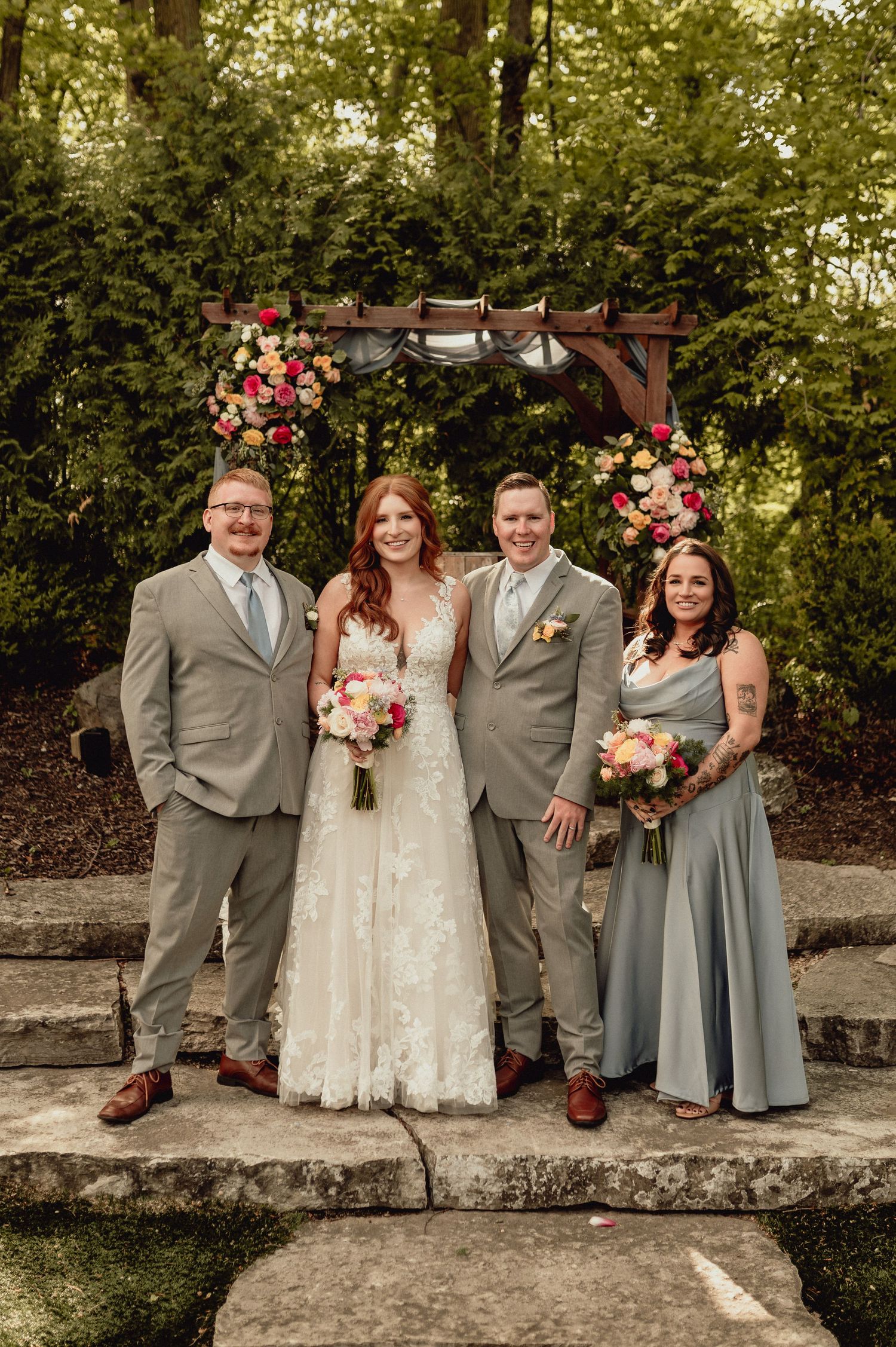 Wedding party in gray suits and dusty blue dresses pose together with pink and white floral bouquets in garden setting.