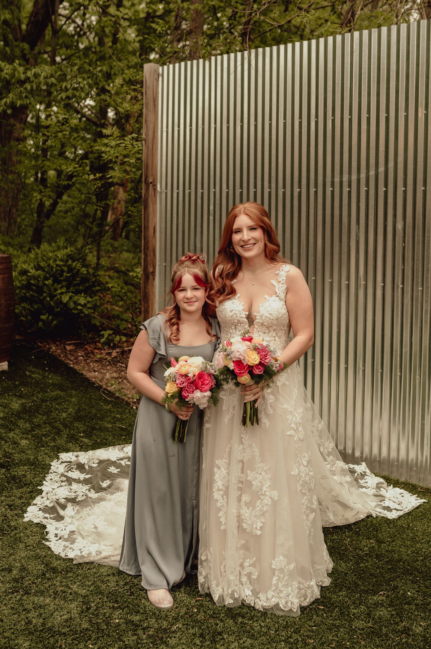 A bride in a lace gown and bridesmaid in gray pose together outdoors against a corrugated metal wall.