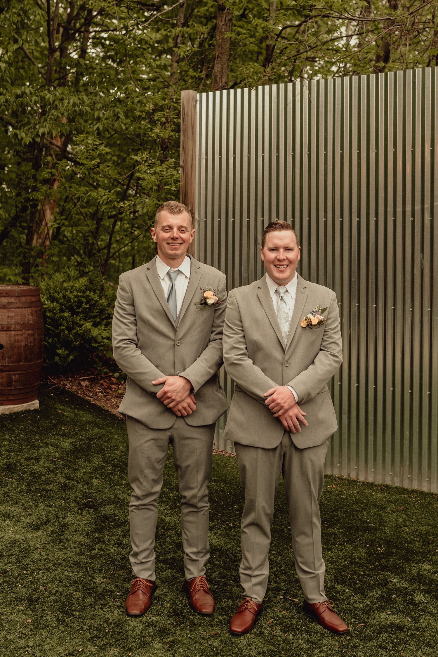 Two groomsmen in light gray suits and brown shoes pose together on grass in front of a metal wall.