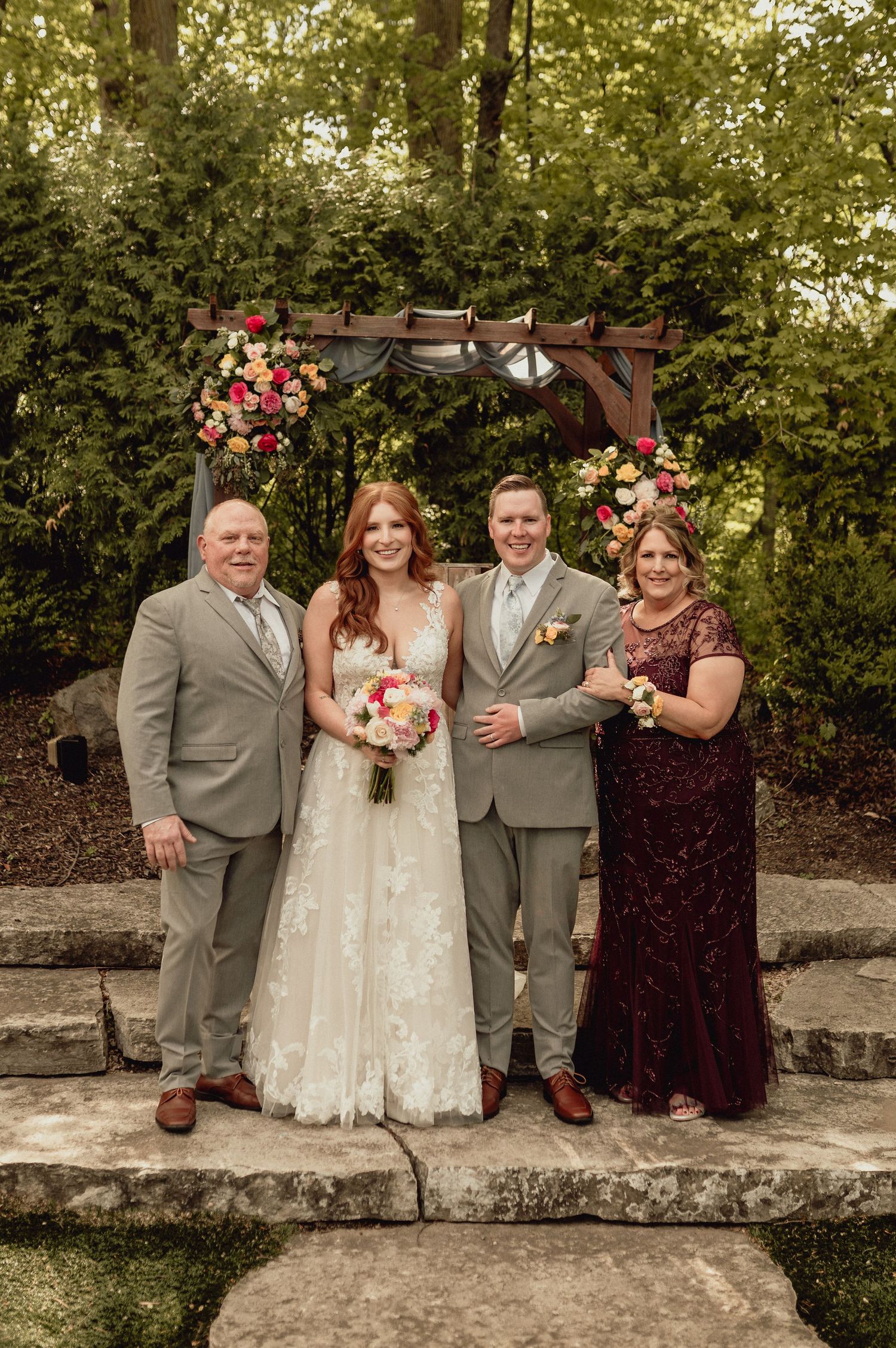 Wedding party in gray and burgundy attire poses together on stone wall surrounded by lush greenery.