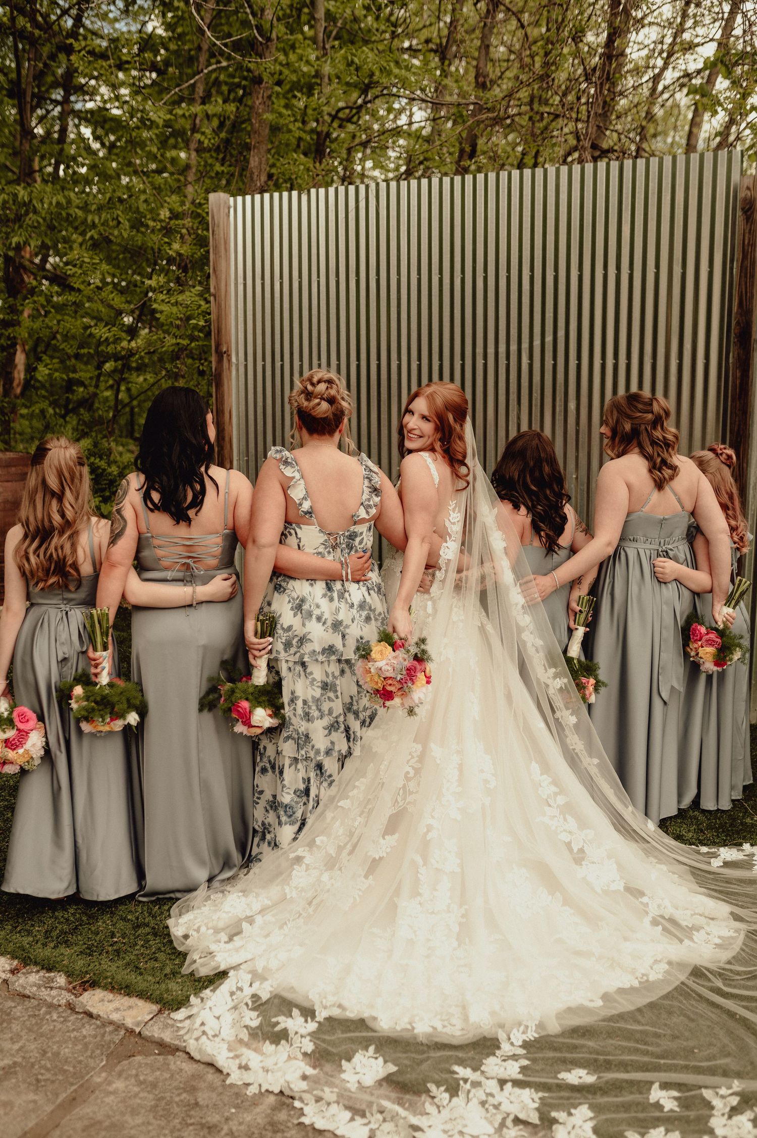 A wedding party of bridesmaids in gray dresses and floral prints stand in a row behind a bride with a flowing lace train.
