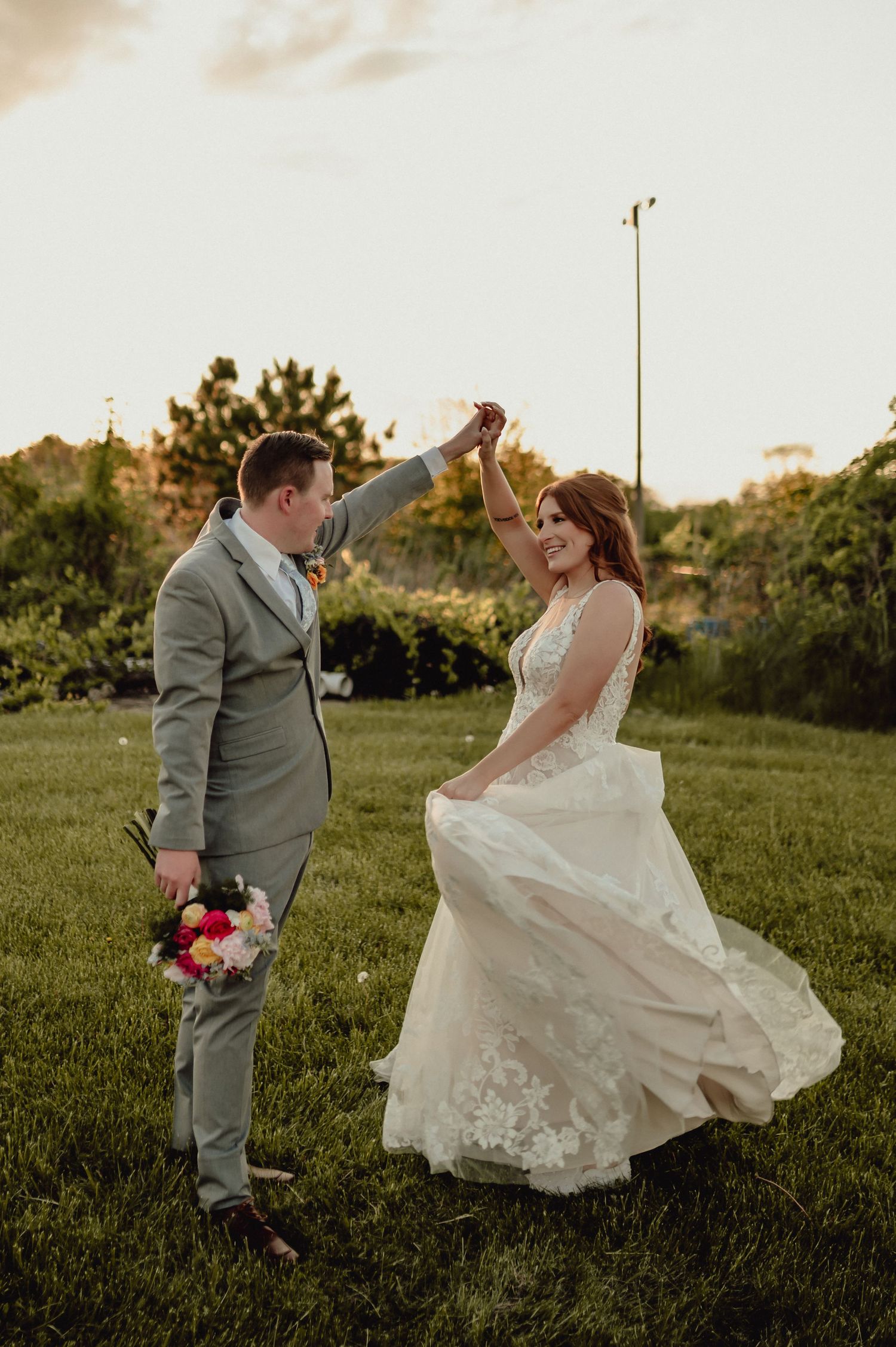 A couple in wedding attire playfully dances together in a grassy field at sunset.