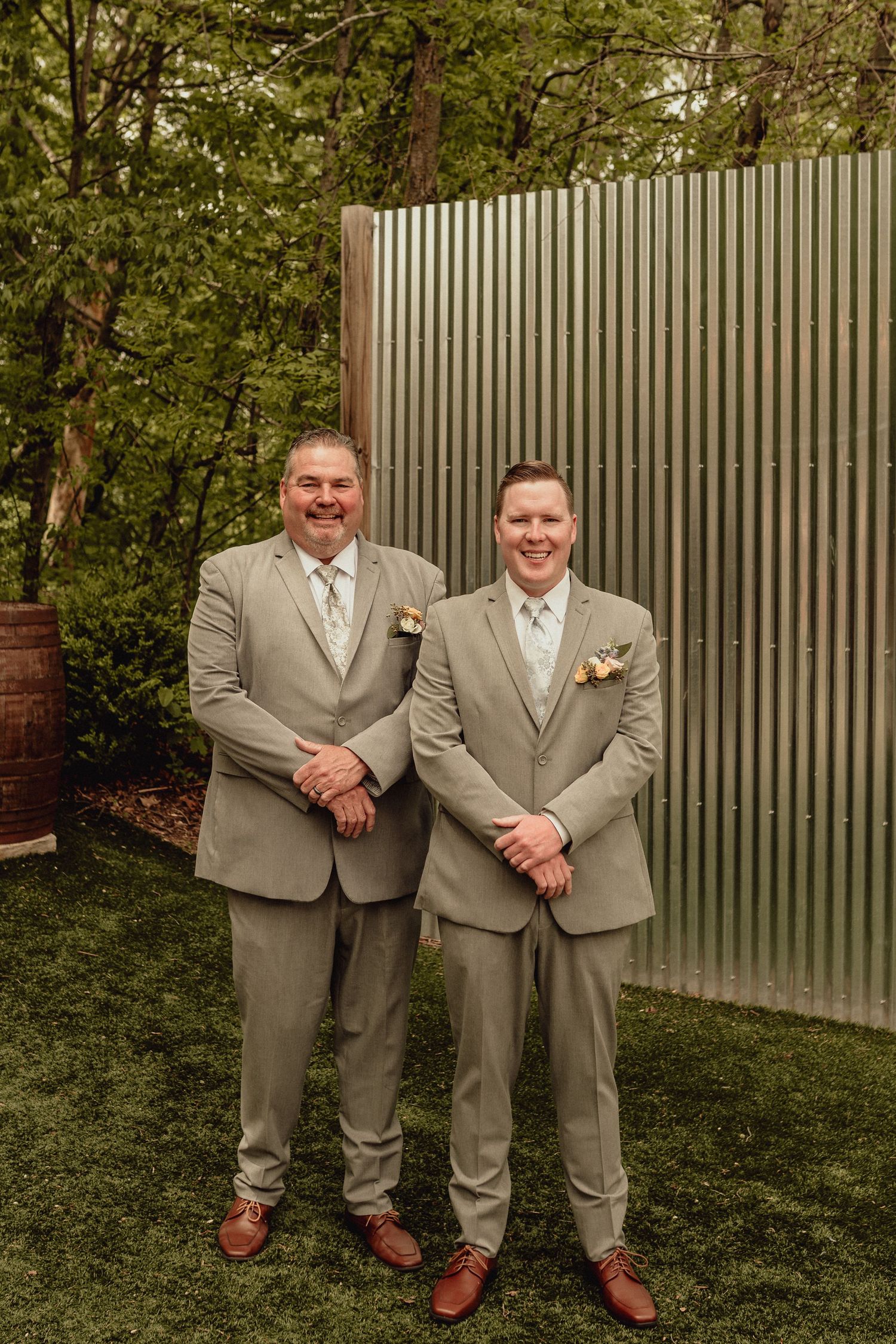 Two people in tan suits and ties stand together on grass near a white fence in five similar poses.