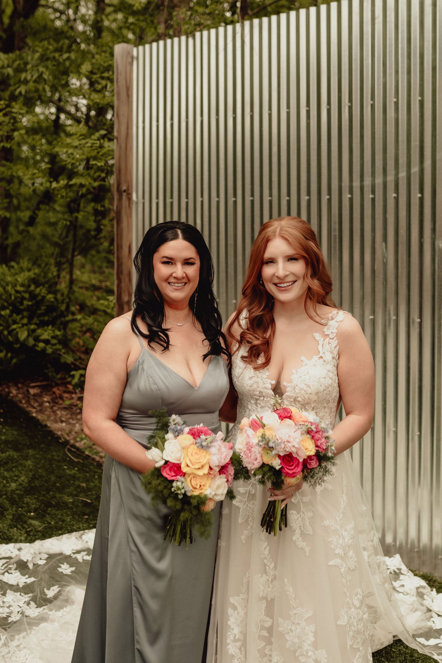 Two women in gray bridesmaid dress and white wedding gown pose together with pink and white floral bouquets.