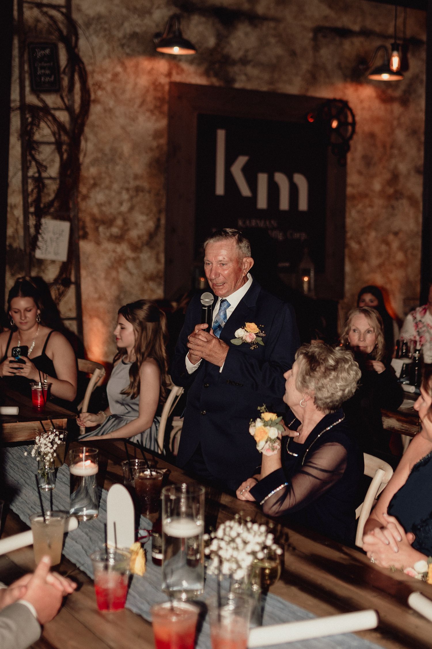 Wedding reception guests gather at a rustic venue with exposed brick walls and warm lighting during dinner speeches.