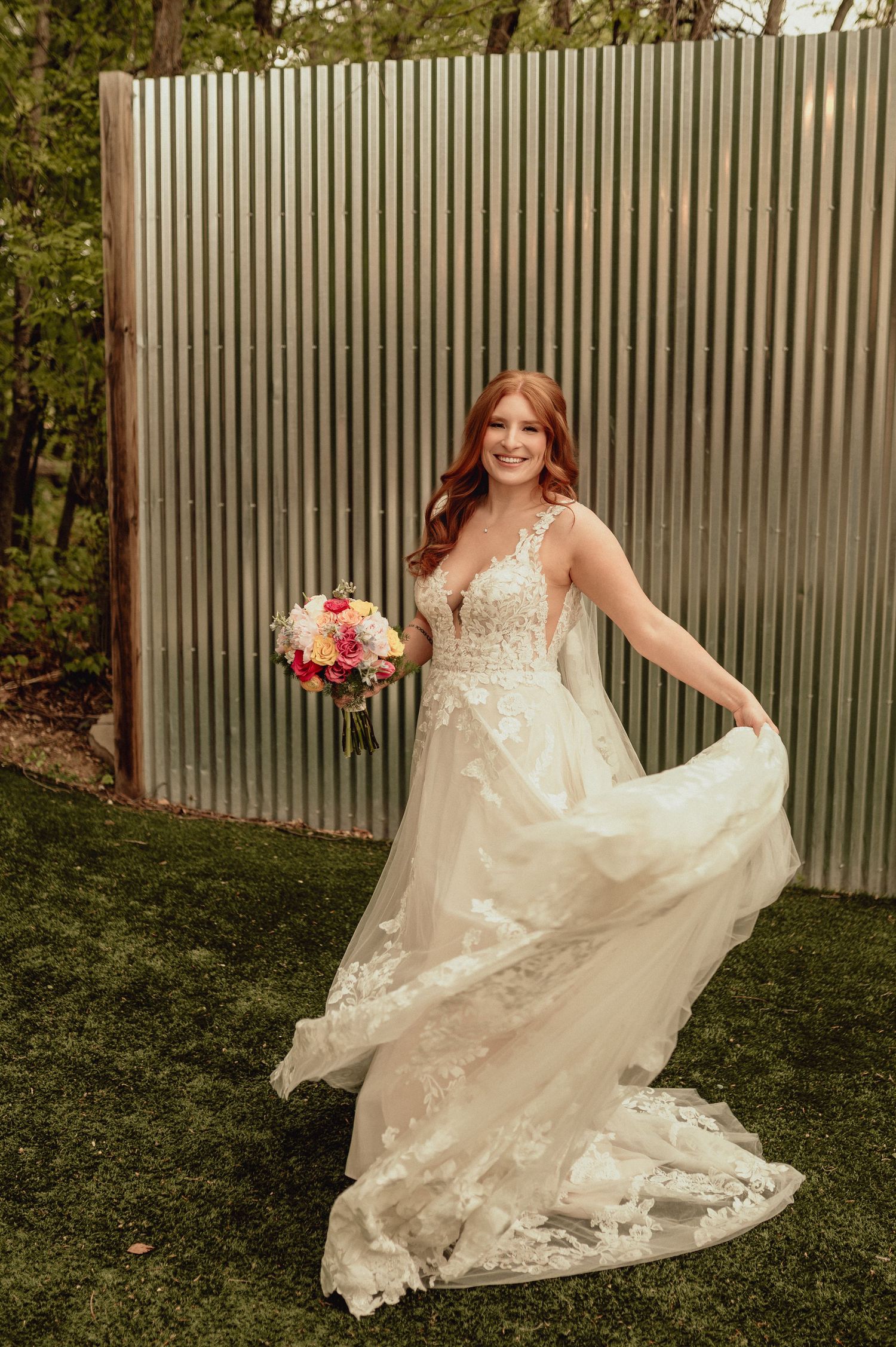 A bride in a flowing lace wedding dress twirls playfully on grass in front of a corrugated metal wall.