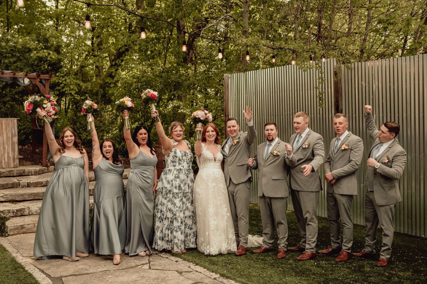 Wedding party in gray attire poses playfully against a wooden fence in a garden setting.