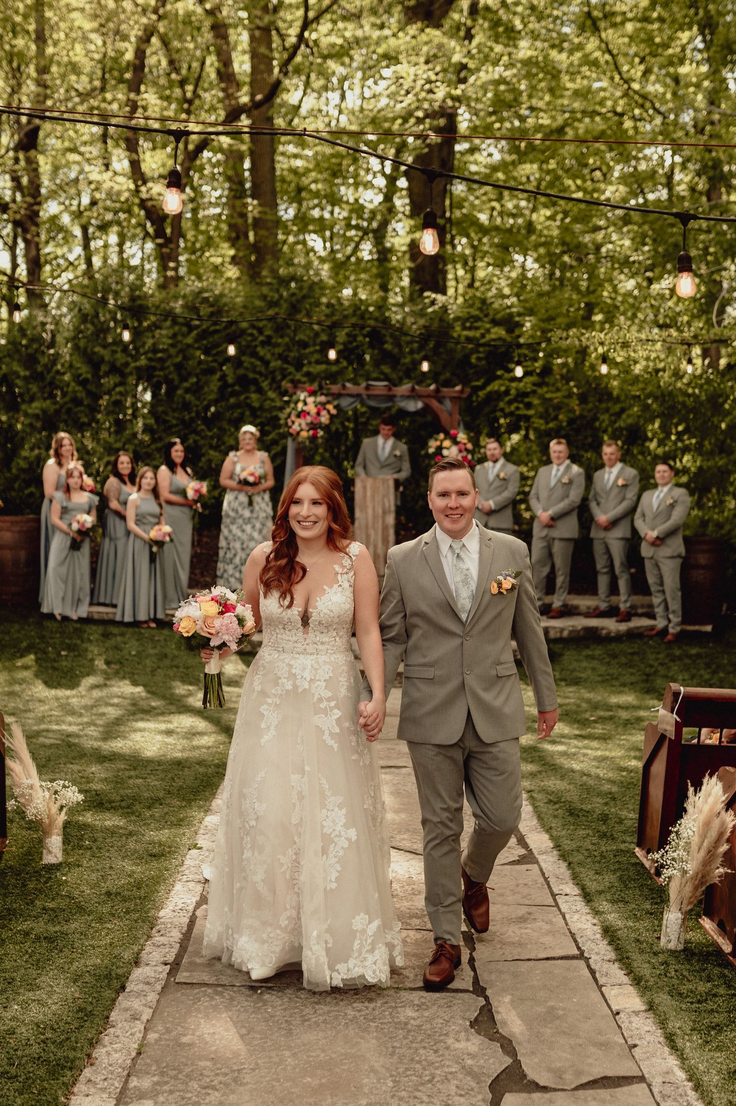 Wedding party poses on stone path in outdoor garden setting with string lights and greenery.