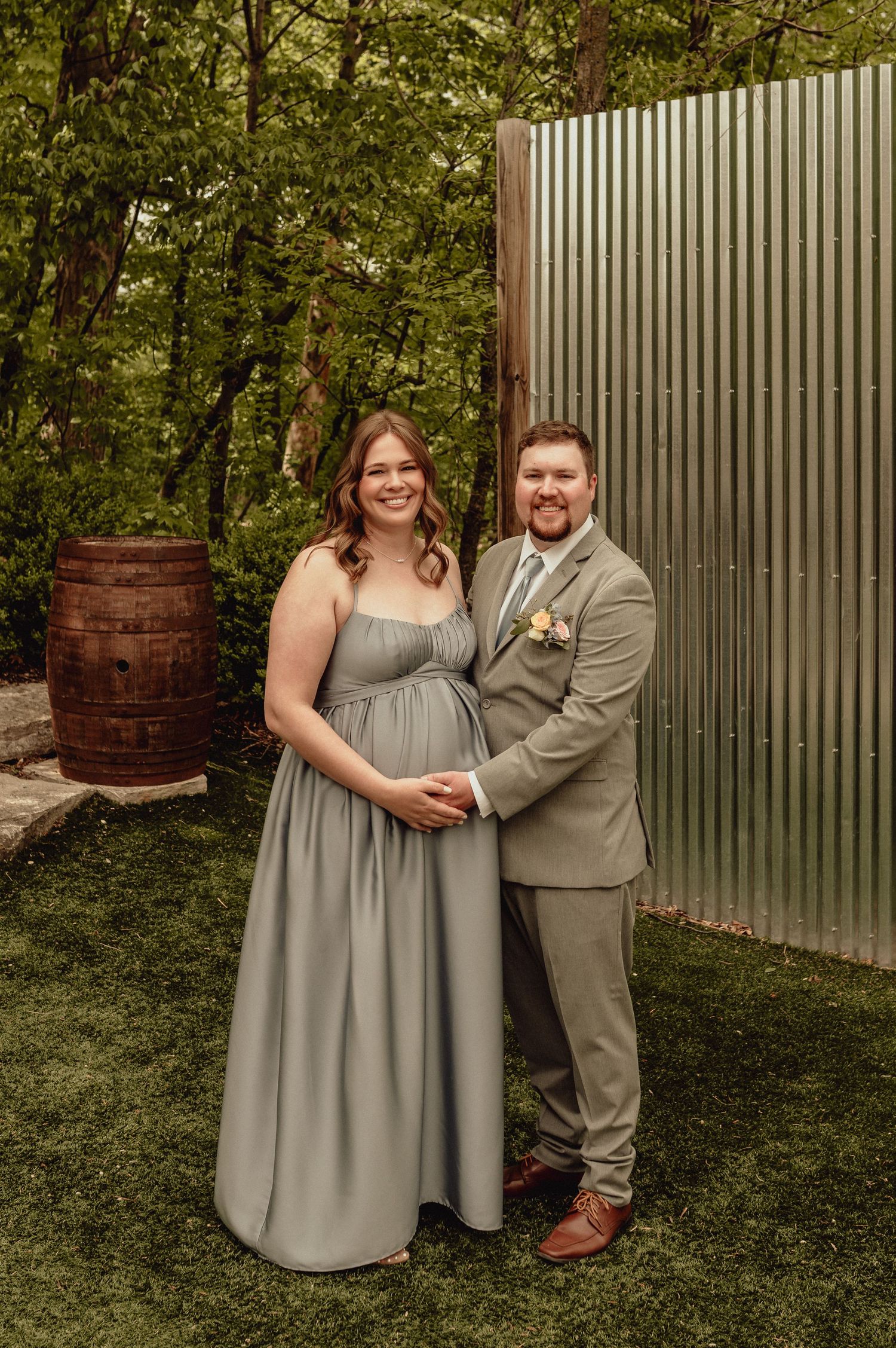 A couple in formal attire poses together in a garden setting with a rustic wooden barrel and metal wall backdrop.