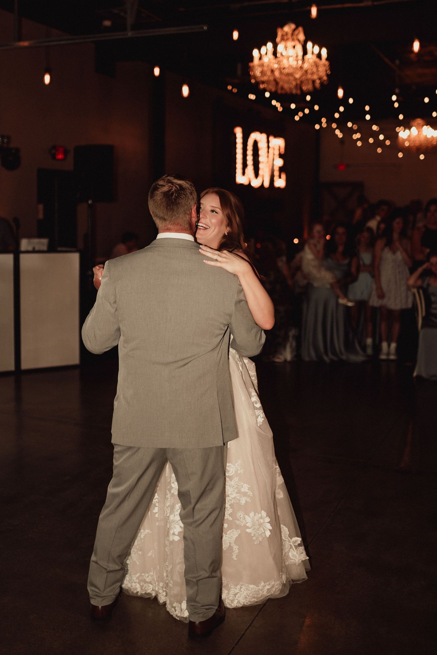 A couple shares a romantic dance moment under a chandelier and string lights at their wedding reception.