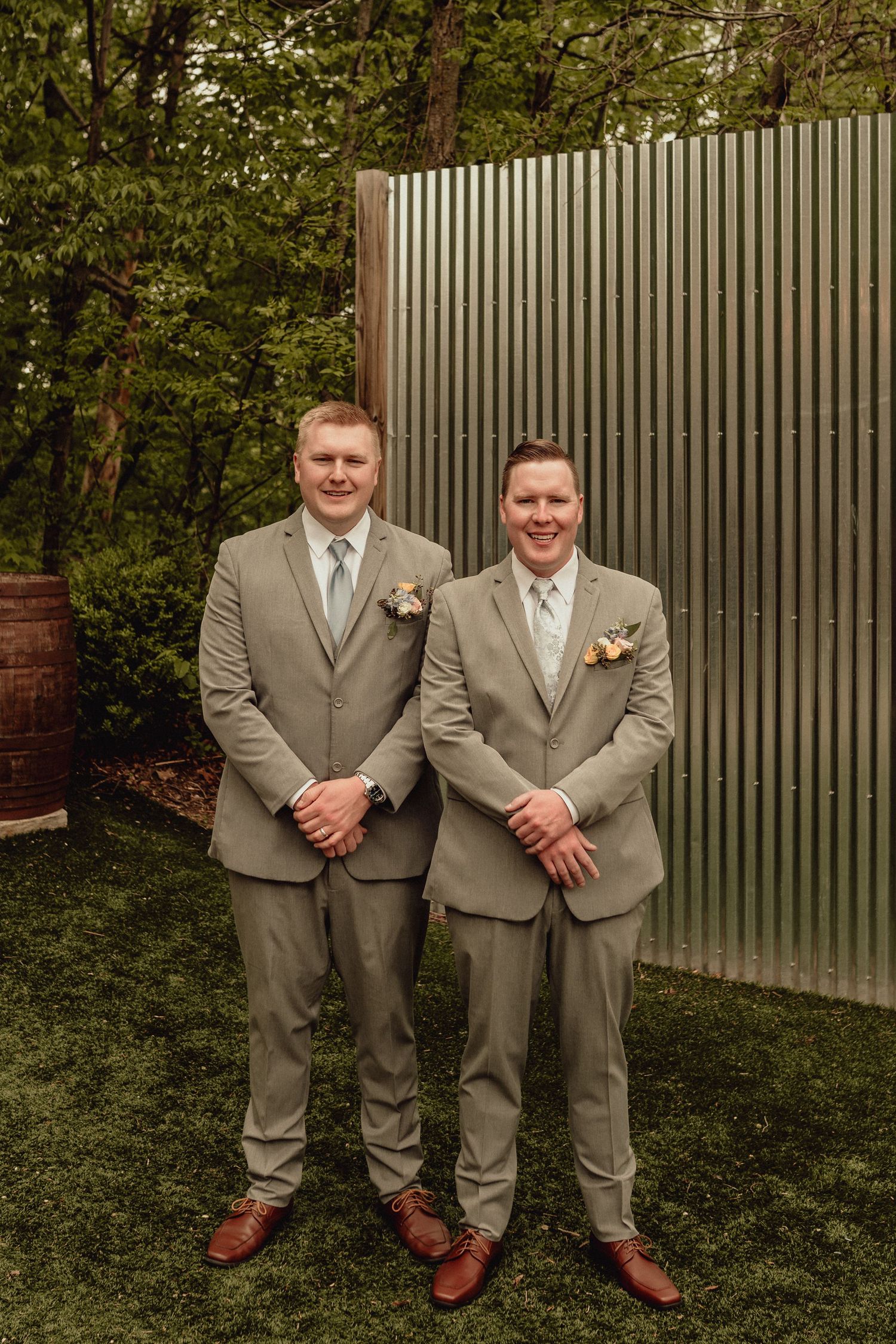 Two men in matching tan suits and brown shoes pose together on grass against a slatted wall in five similar photos.
