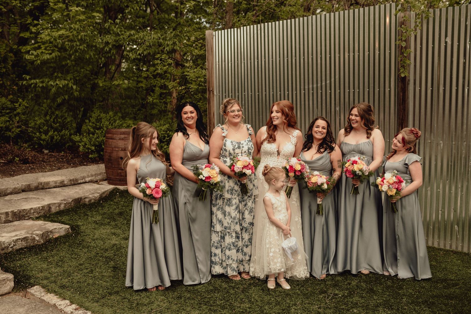 A bridal party in flowing gray dresses and floral prints poses together on a lawn with bouquets against a modern wall backdrop.