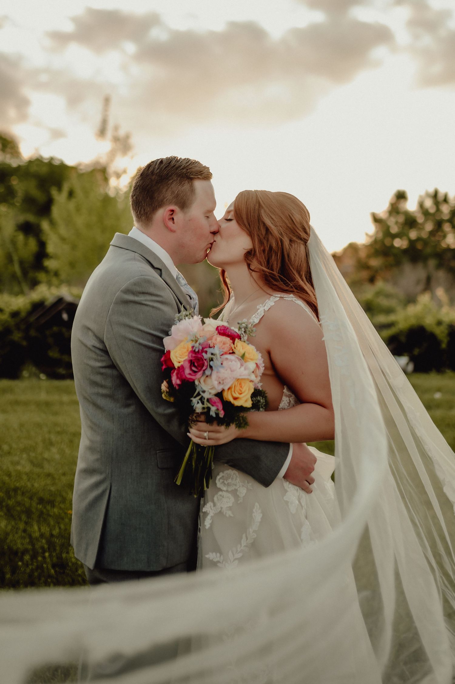 A romantic sunset wedding portrait shows a couple embracing while holding a vibrant bouquet of flowers.