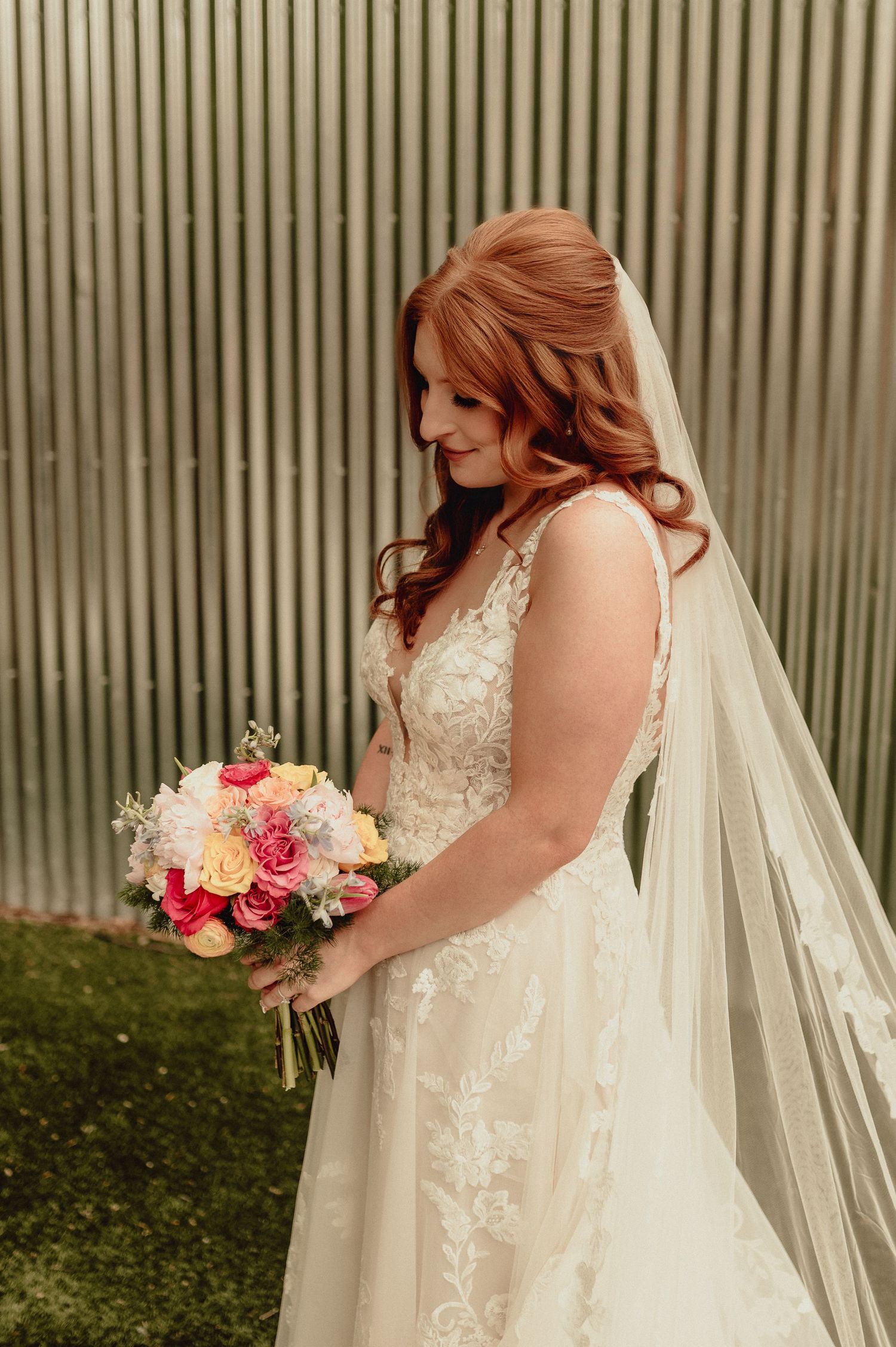A bride in a lace wedding gown with flowing veil holds a pink and coral bouquet against a striped wall backdrop.