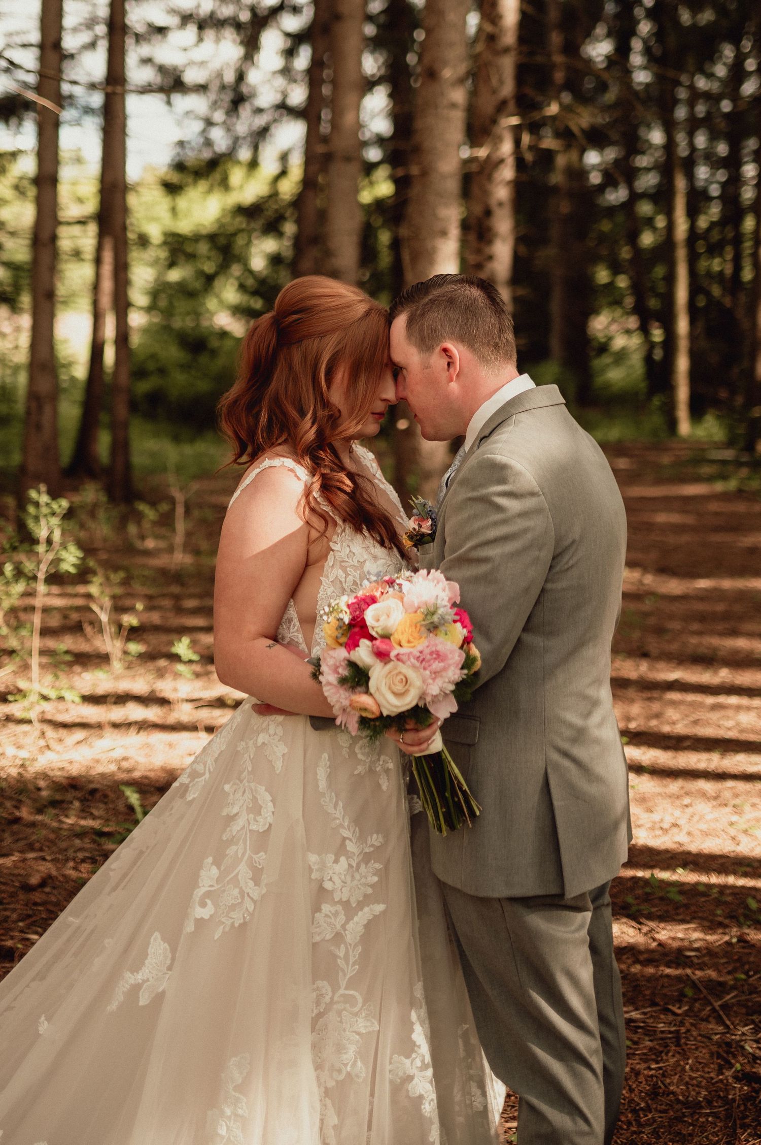 Romantic wedding couple embraces in a sunlit forest setting with a beautiful pink and white floral bouquet.