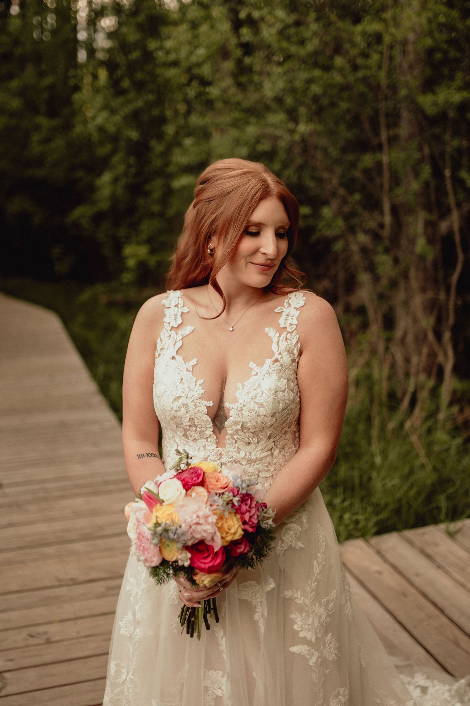 Bride in lace wedding dress holds vibrant pink and purple floral bouquet while standing on wooden boardwalk.