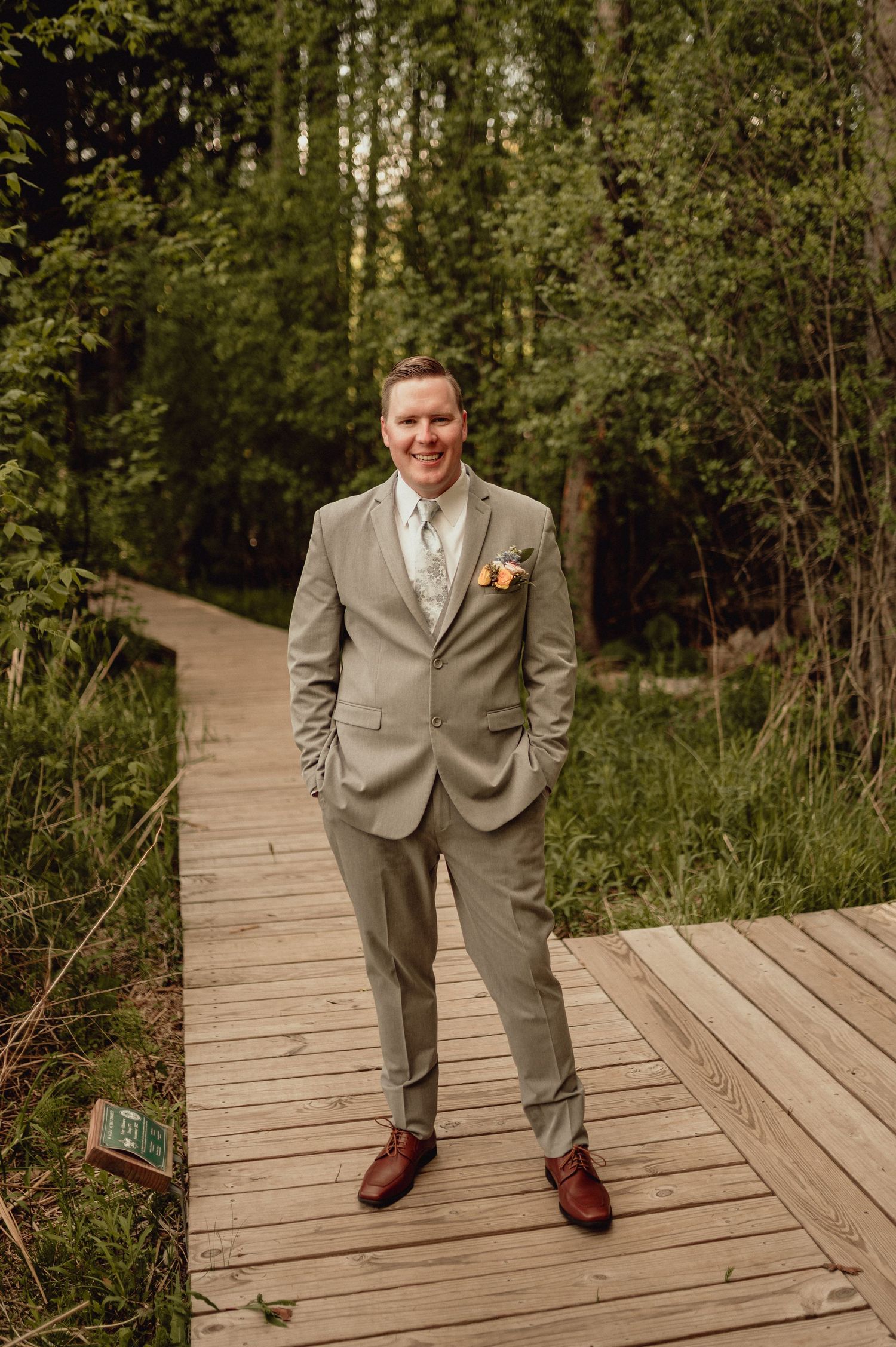 A groom in a light gray suit and burgundy shoes stands on a wooden boardwalk path surrounded by lush forest greenery.